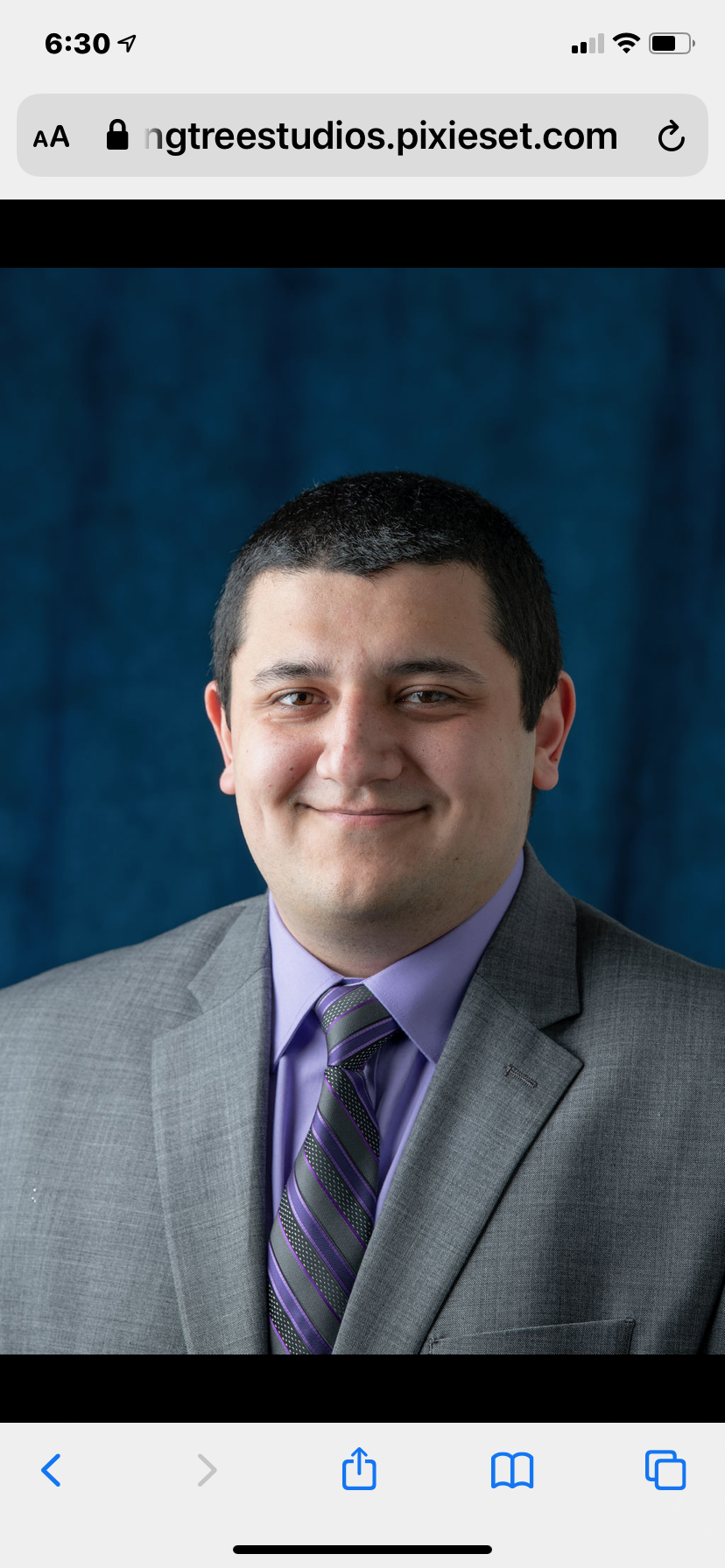 A professional headshot of a man in a gray suit, purple shirt, and striped purple and gray tie, smiling against a blue textured backdrop.