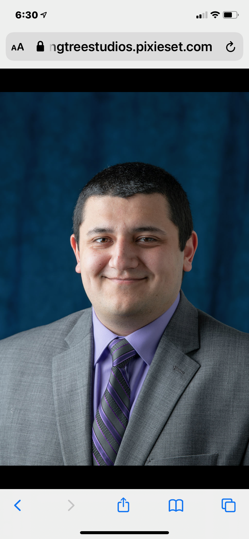Professional headshot of a man with short dark hair, wearing a gray suit, purple shirt, and striped purple and black tie, set against a blue background.