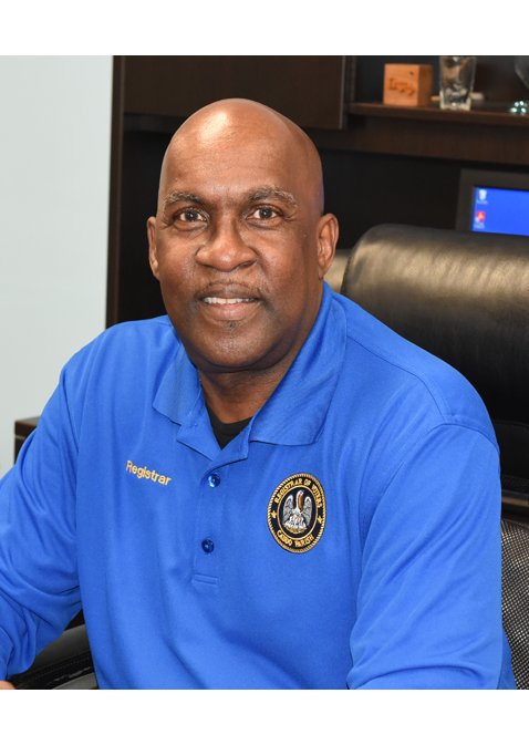 A smiling man sitting in an office, wearing a blue shirt with a 'Registrar' name tag and an official emblem on the chest.