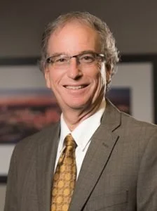 A middle-aged man with glasses, gray hair, wearing a gray suit, white shirt, and a patterned gold tie, smiling in an office setting.
