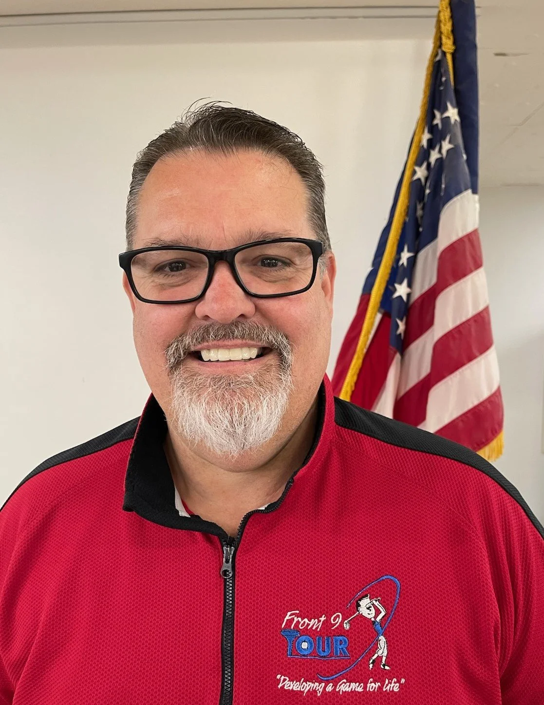 A man with glasses, a beard, and a mustache smiling in front of an American flag. He's wearing a red sports jacket with the logo 'Front 9 Tour' and the slogan 'Developing a game for life.'