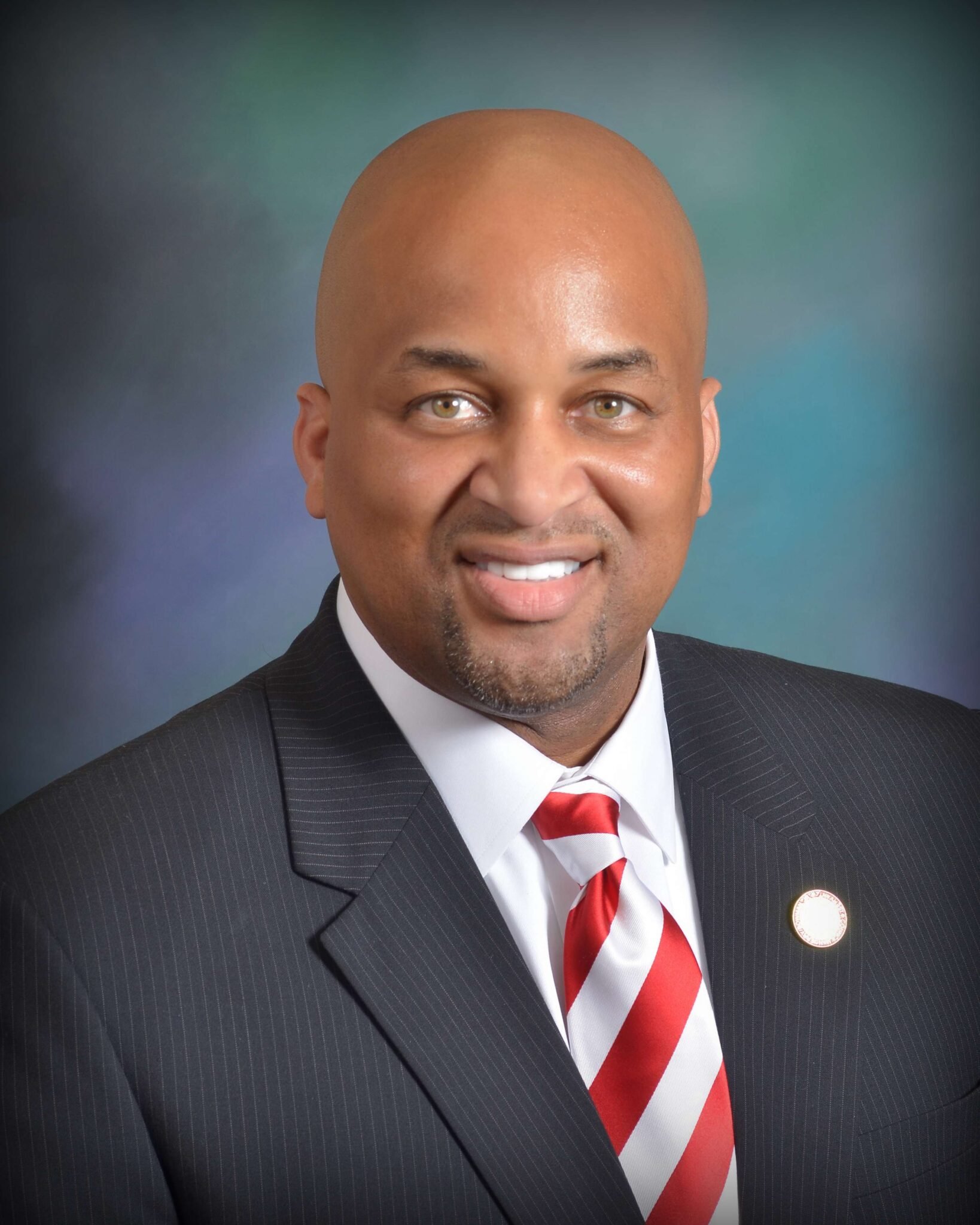 A man wearing a dark pinstripe suit, white shirt, and red and white striped tie, smiling in a professional portrait with a blue-green gradient background.