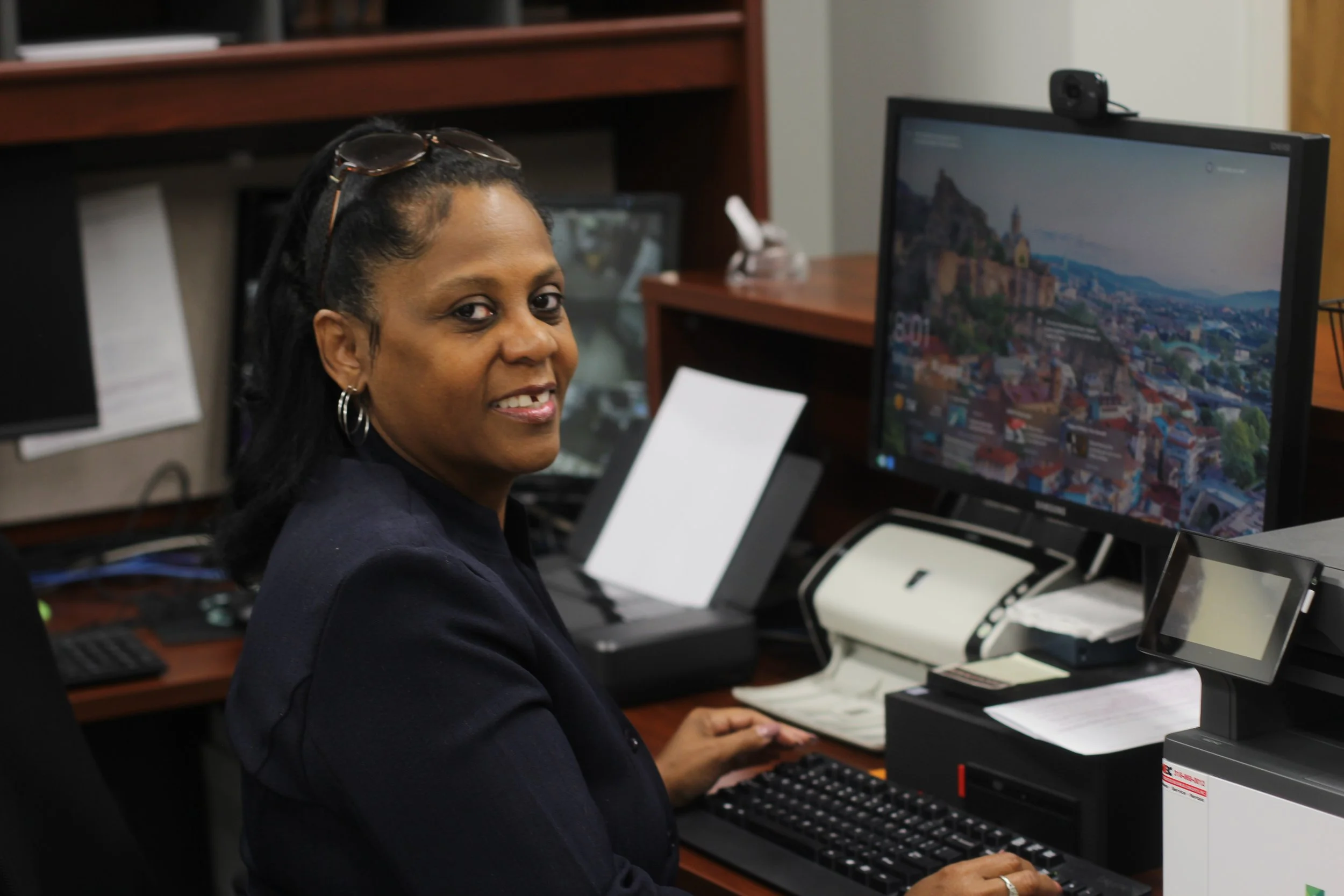 A smiling Black man in a blue polo shirt sitting at a desk in an office.