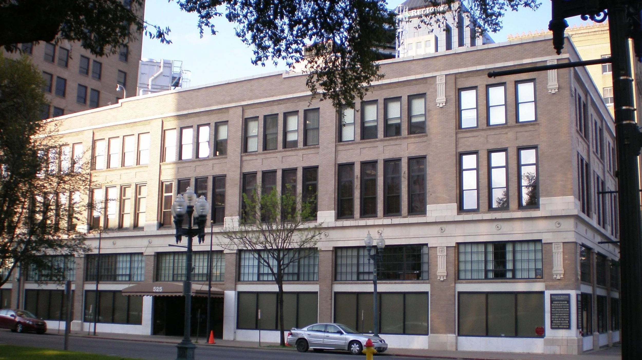 A multi-story brick building with large windows, street lamps, parked cars, trees, and a sidewalk in front. Part of the cityscape visible in the background.