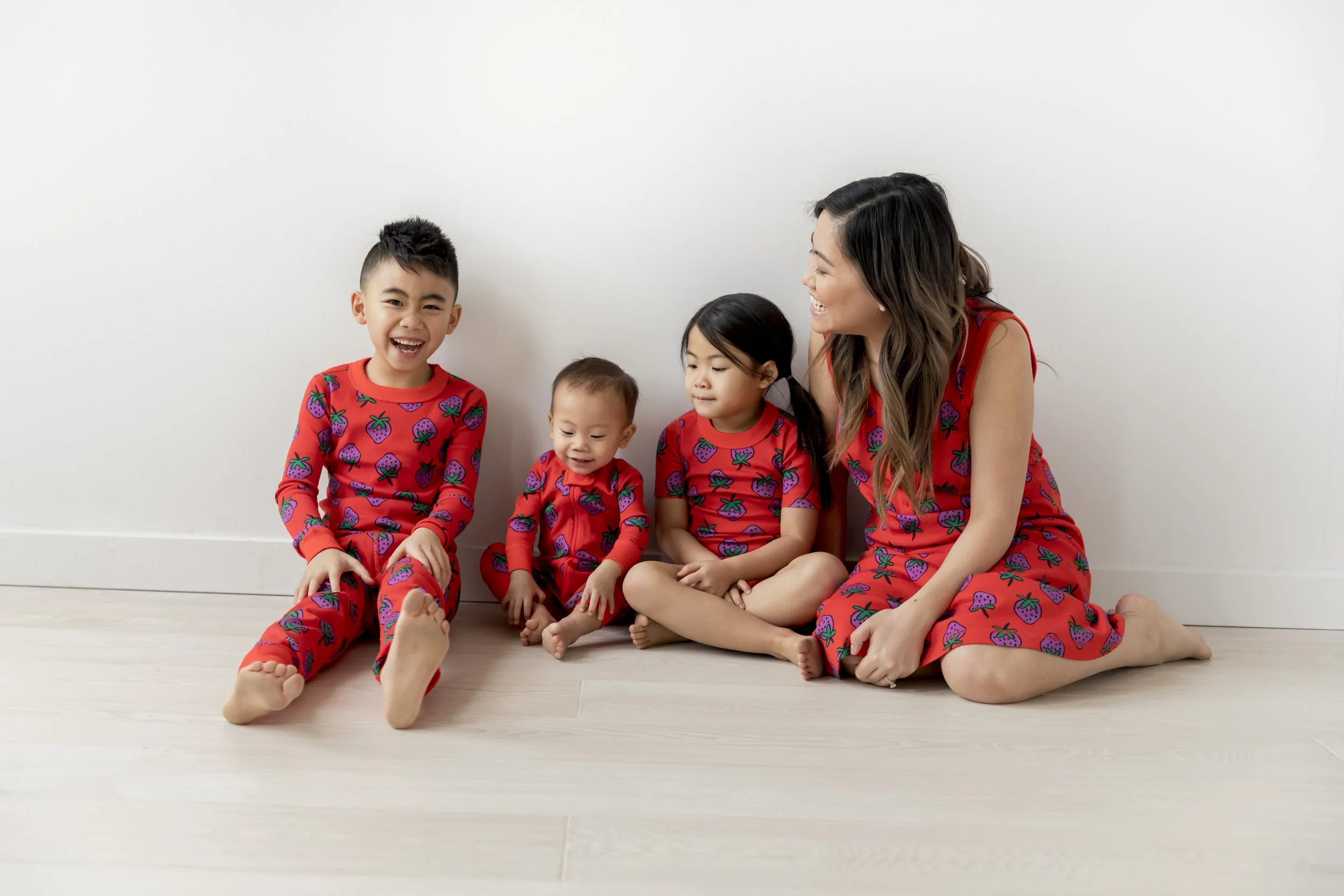 A woman and four children sitting on the floor against a white wall, all wearing matching red pajamas with purple strawberries prints and smiling or looking content.