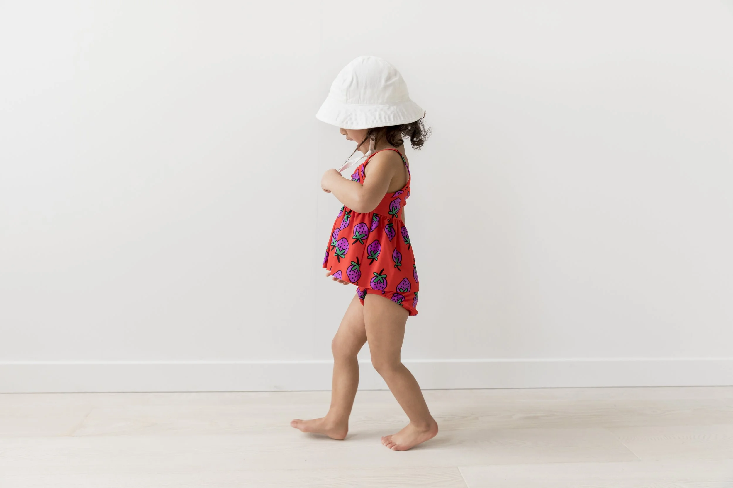 A young girl with curly hair wearing a white hat, a colorful strawberry-patterned dress, and shorts, standing barefoot on a light-colored wooden floor against a plain white wall.