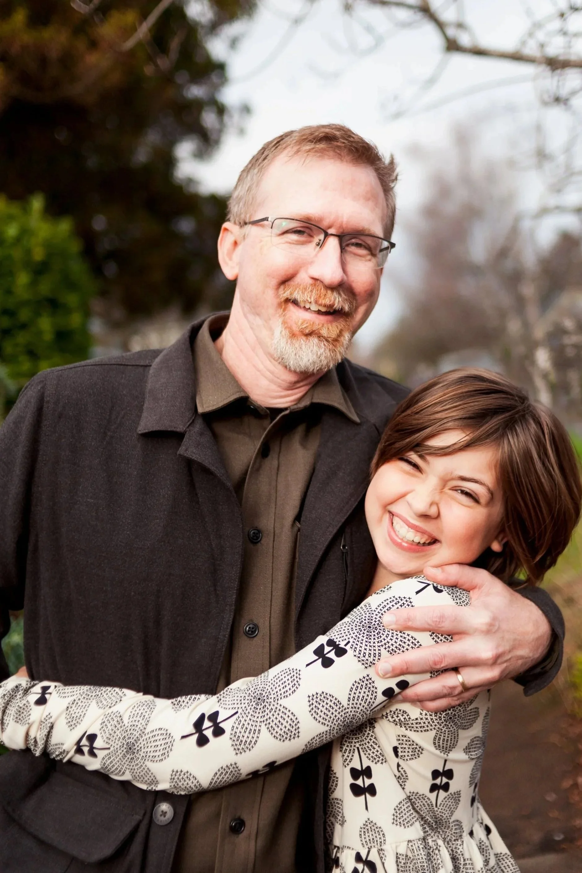 A man with glasses and a beard smiling, standing outdoors with a woman who is hugging him, in a park or garden with trees in the background.