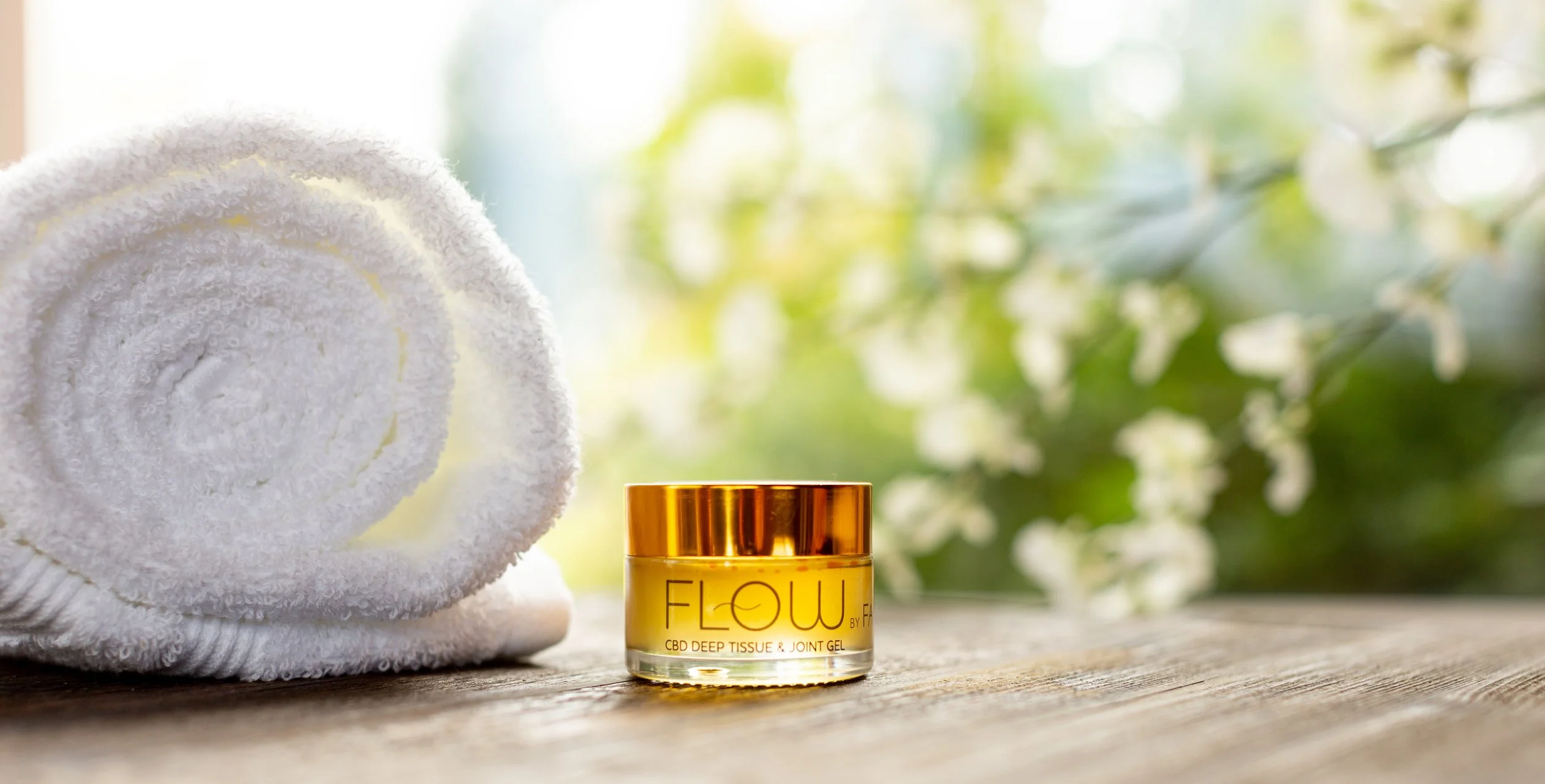 A rolled white towel, a yellow jar of CBD deep tissue and joint gel with a gold lid, and a blurred background of green leaves and white flowers.