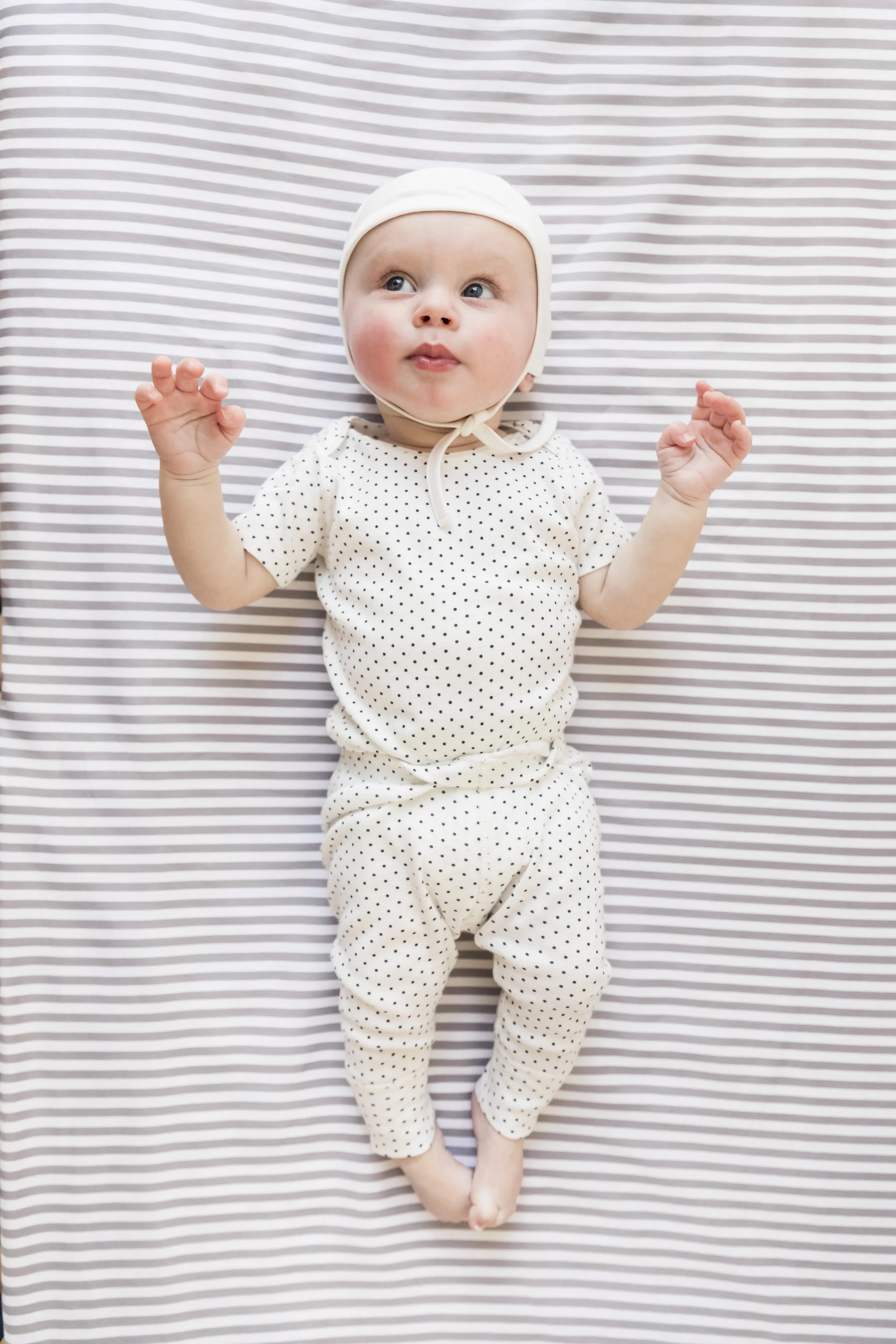 A baby lying on a striped bedsheet wearing a white outfit with black polka dots and a white bonnet, looking alert with hands raised.