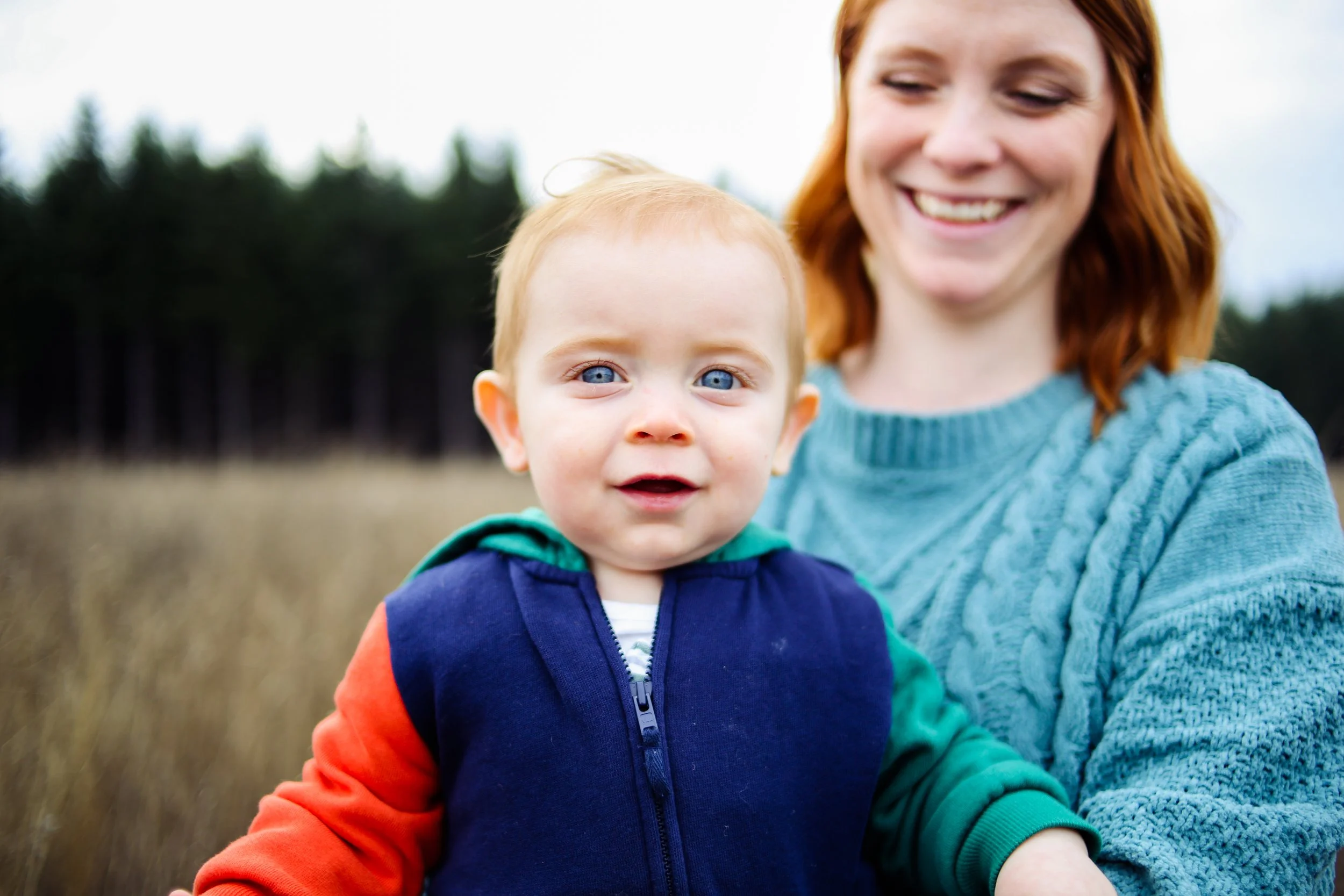A smiling woman with red hair holding a young boy with blue eyes and blonde hair in an outdoor setting with trees in the background.