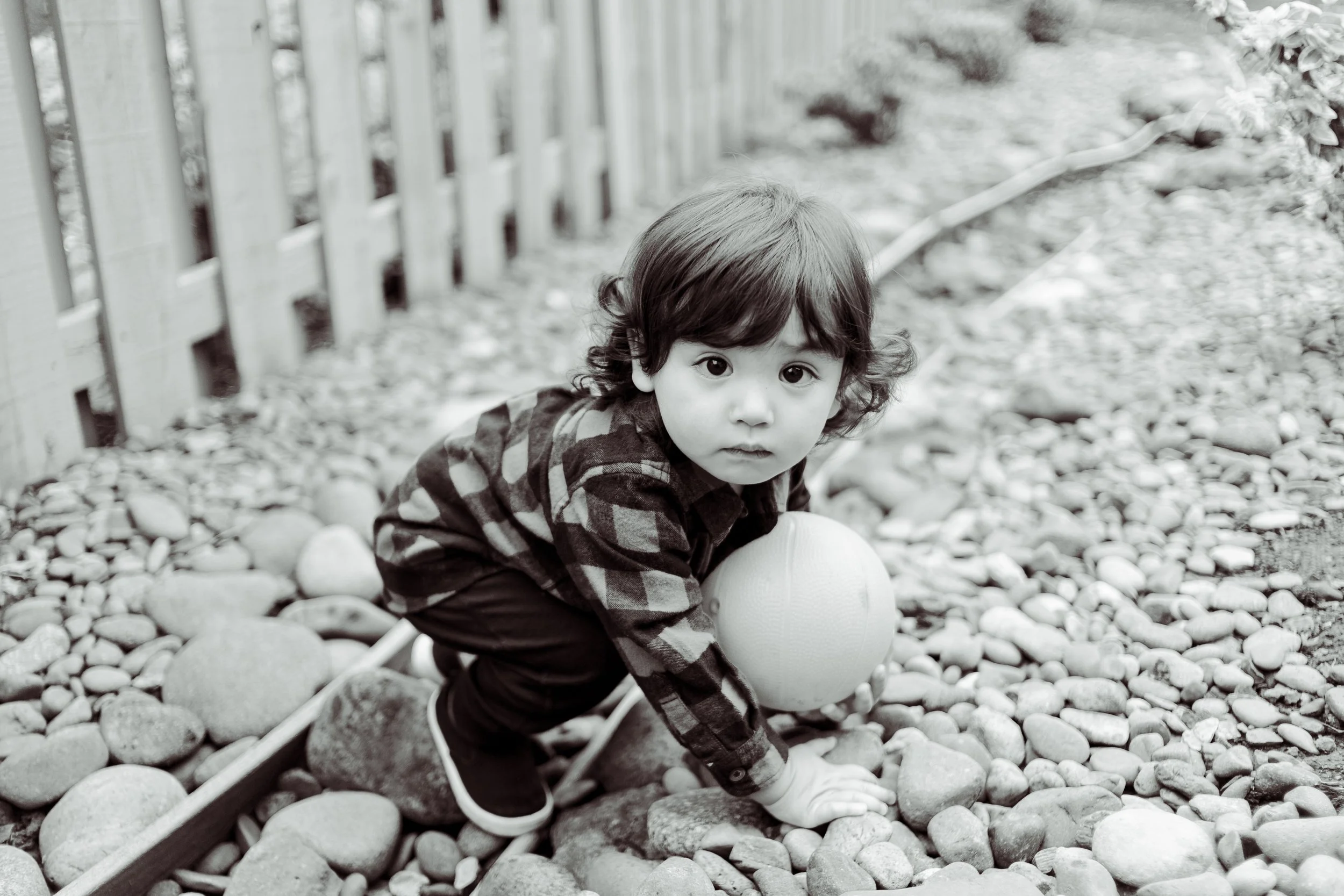 A young boy with dark curly hair and light-colored eyes crouching on a rocky surface outdoors next to a white ball, with a wooden fence in the background.