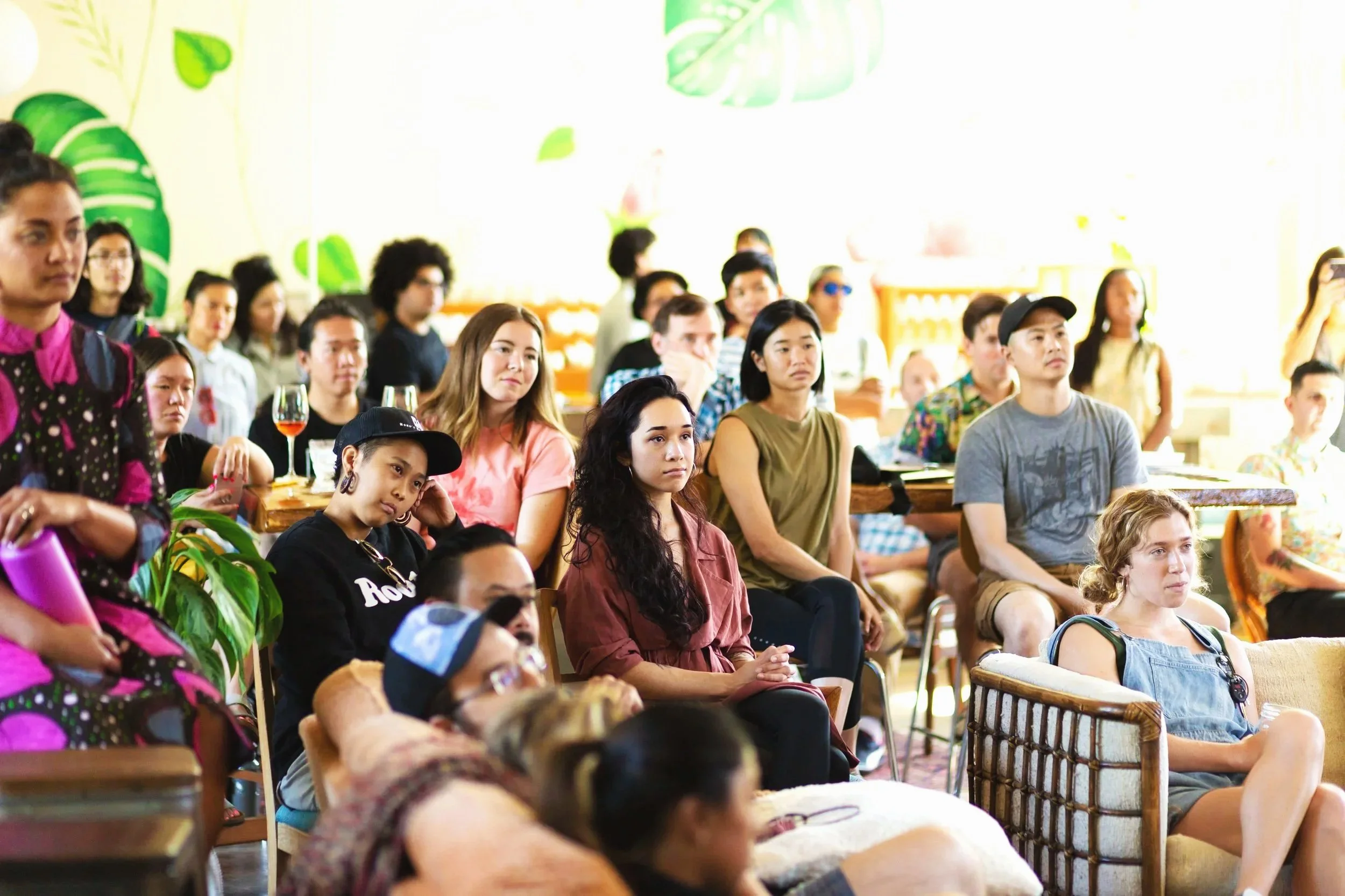 A diverse group of people are seated attentively in a brightly lit indoor setting, possibly attending a presentation or event, with some holding drinks and others listening intently.
