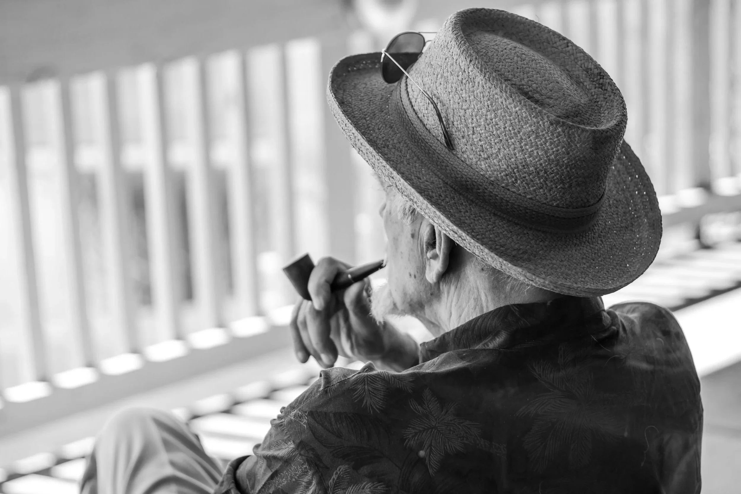 A person wearing a straw hat with sunglasses on top, smoking a pipe, and sitting outdoors.