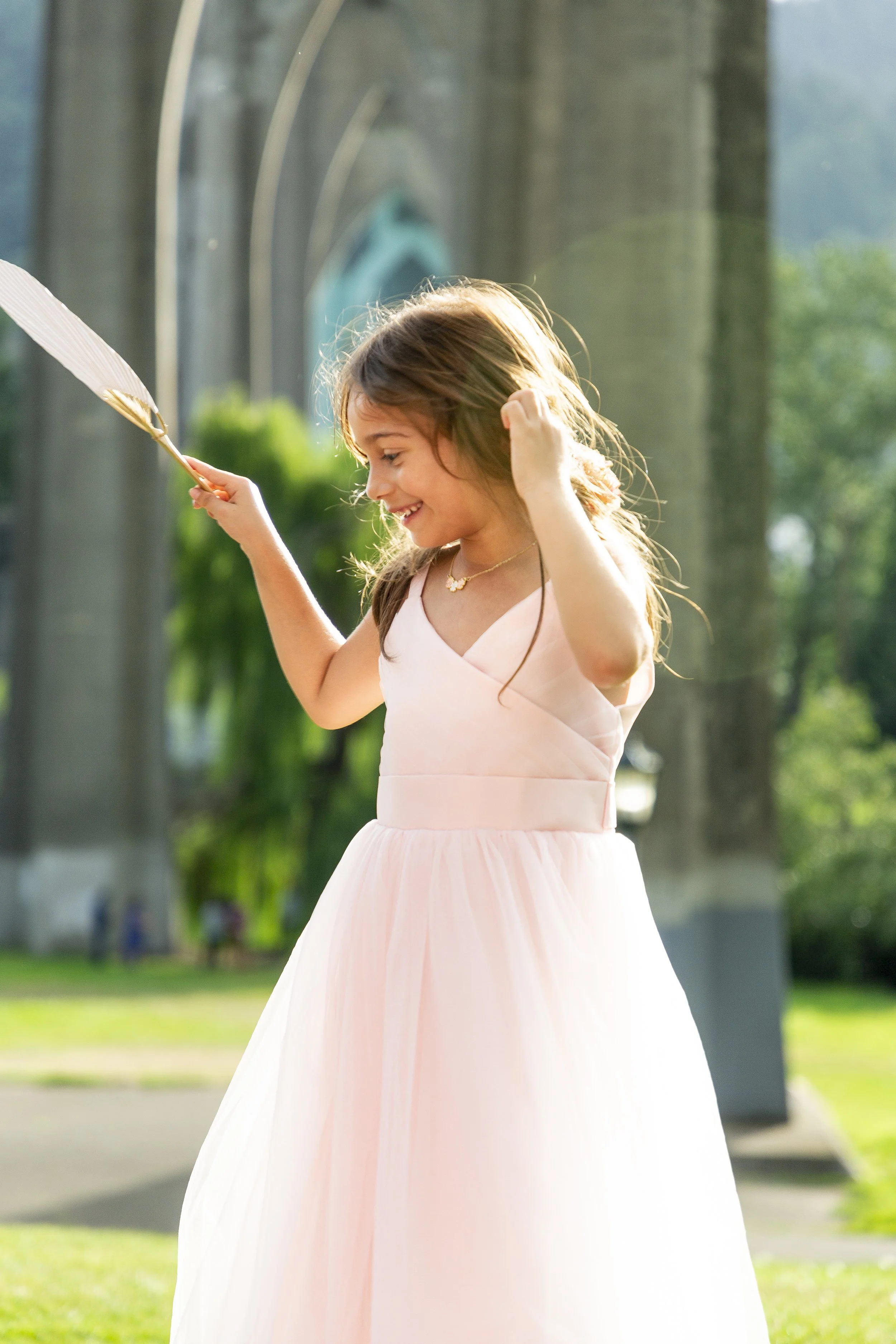 A young girl in a light pink strapless dress playing with a feather in a park with green trees and an arched bridge in the background.