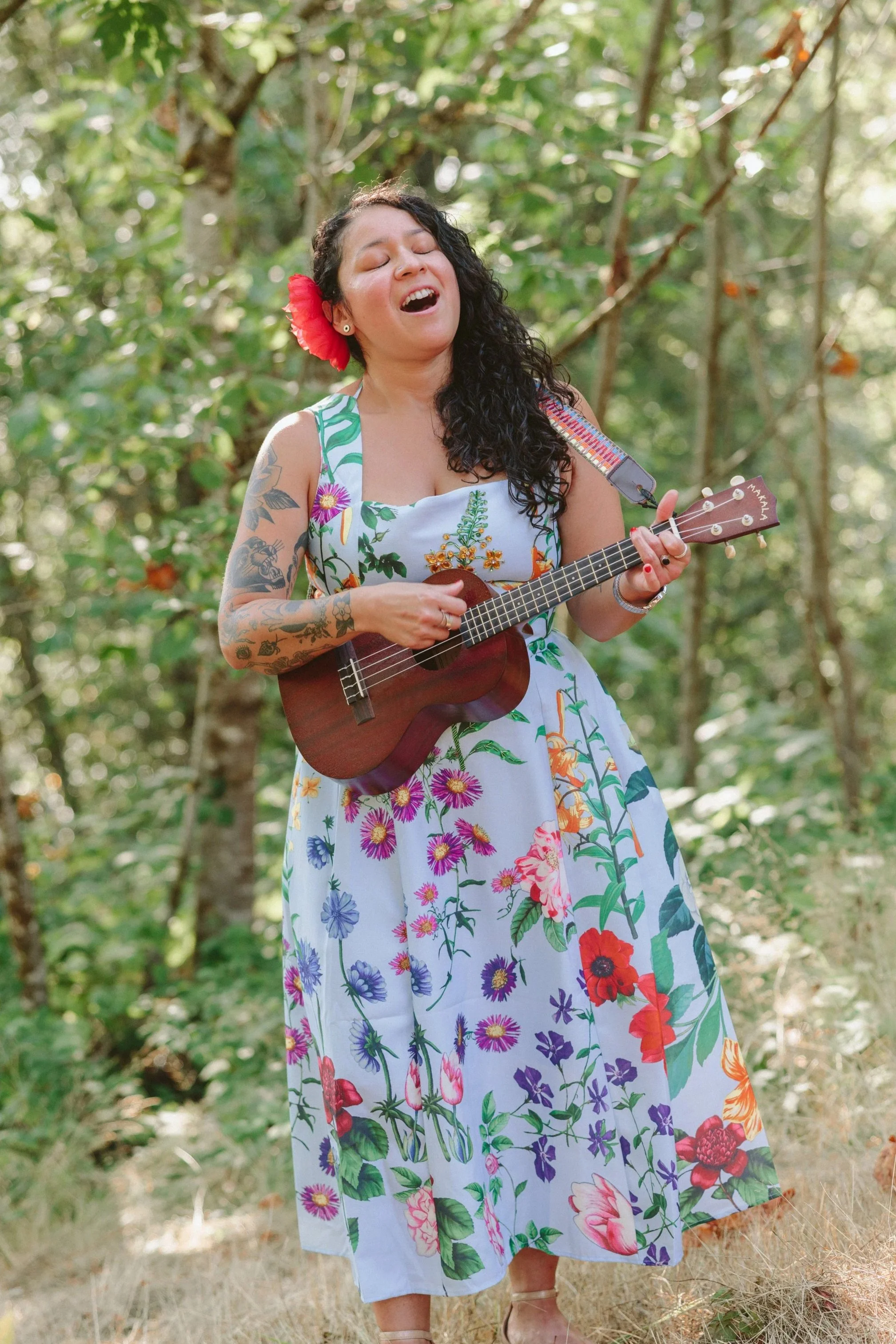 A woman wearing a floral dress and a red flower in her hair is singing and playing a ukulele in a forested area with green trees.