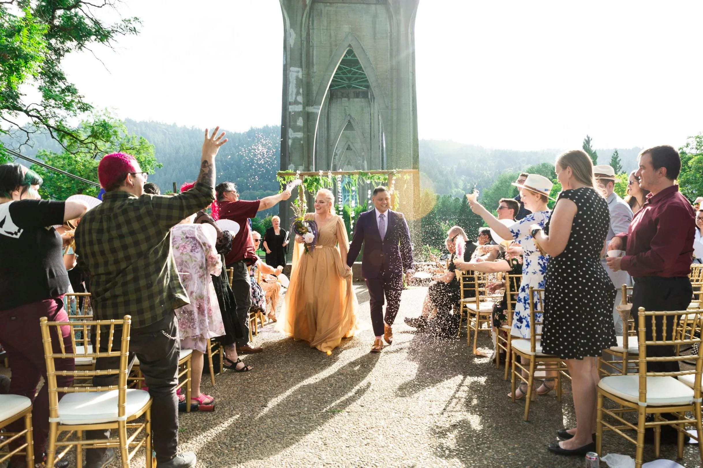 A wedding ceremony outdoors with a crowd of people dressed in semi-formal attire, some standing and others seated, with a bride and groom walking together under a large concrete bridge in the background, as guests throw confetti.