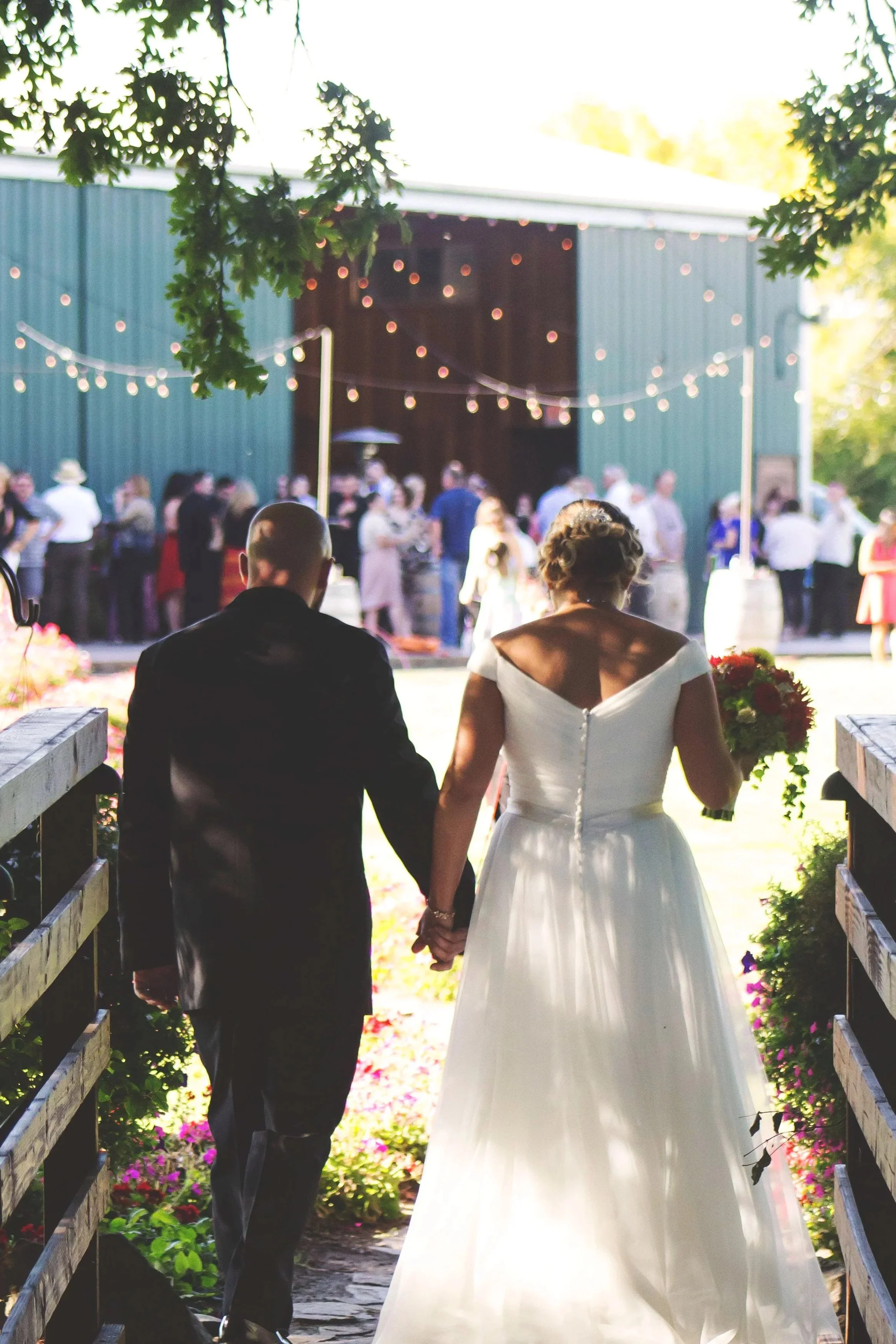 A bride and groom holding hands walking towards a barn decorated with string lights during a wedding reception outside. Guests are gathering in the background.