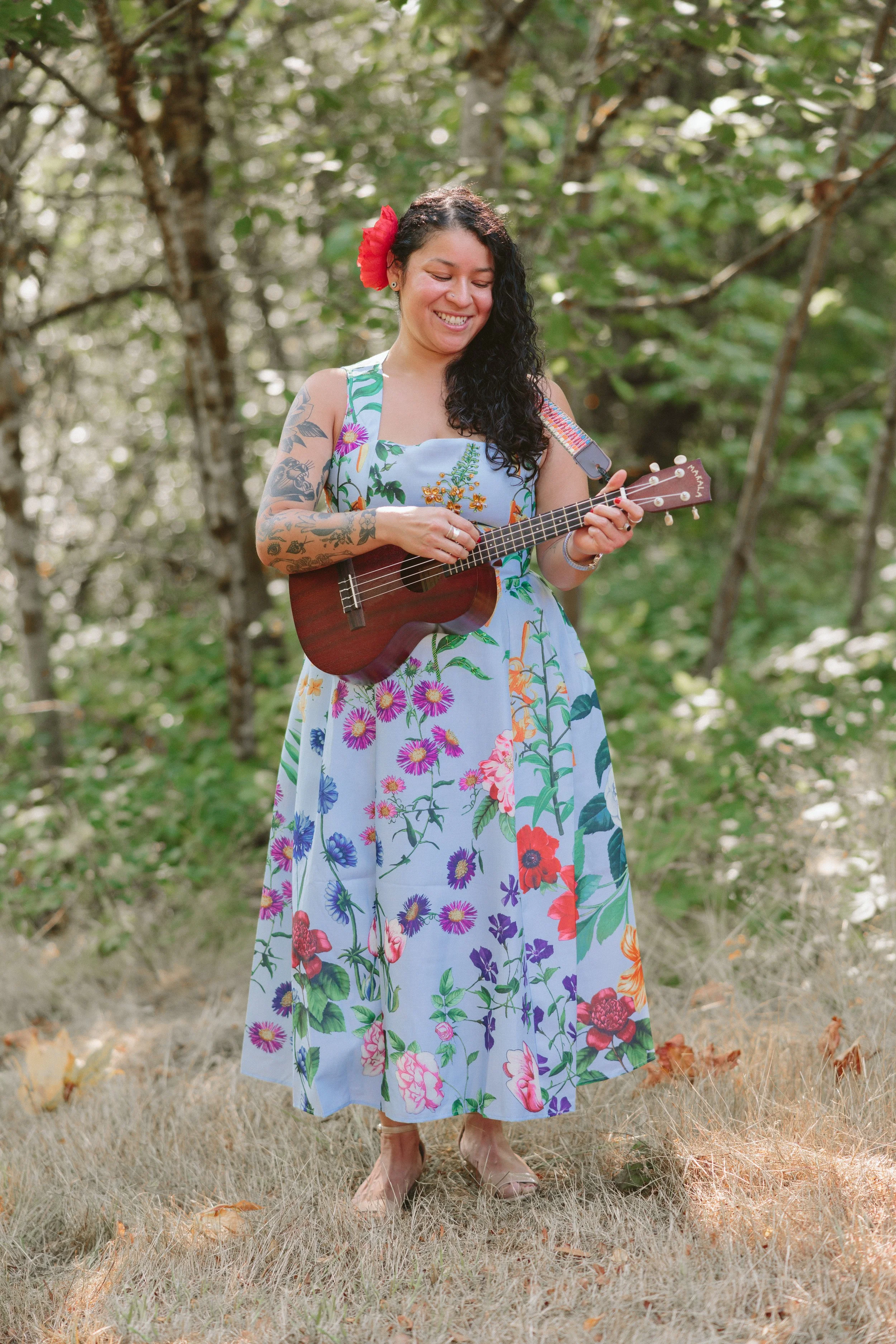 A woman with long, curly black hair, wearing a colorful floral dress, standing outdoors among trees, playing a ukulele with a big red flower in her hair, smiling.
