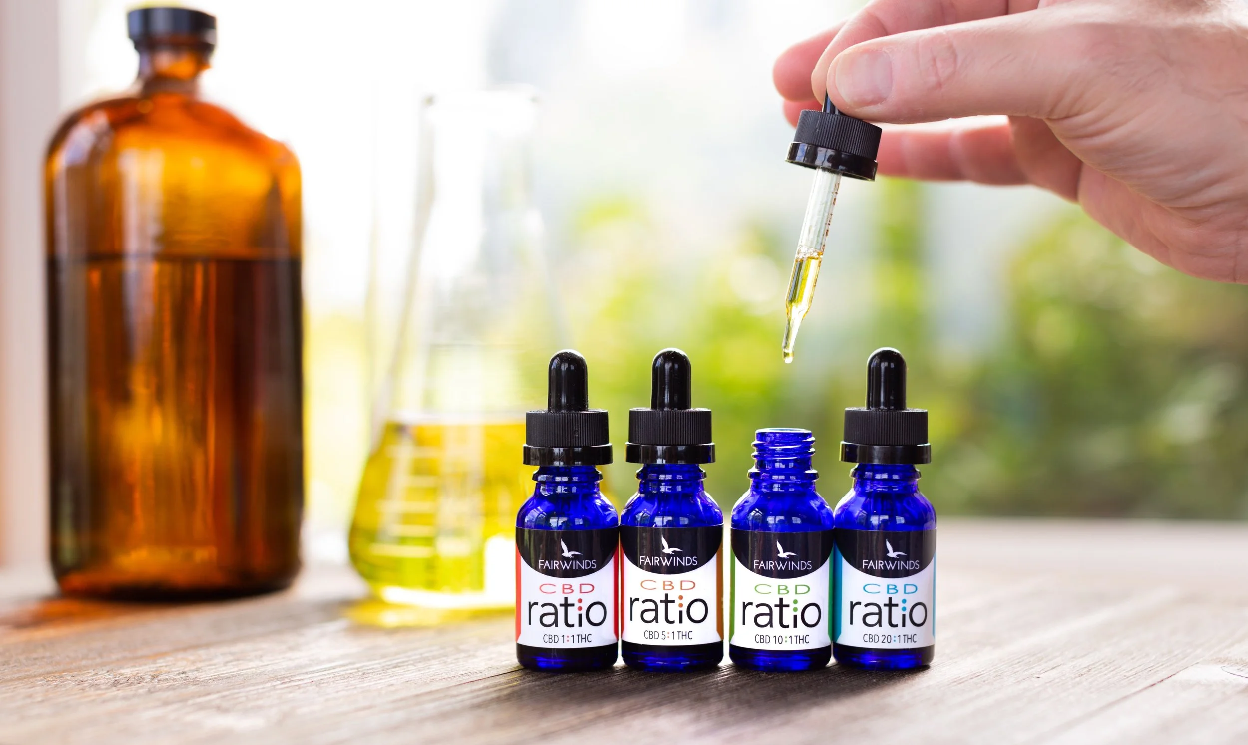 Several small blue bottles of CBD oil with dropper caps, labeled with the brand 'Fairwinds' and various CBD to THC ratios, are arranged on a wooden surface. A hand is holding a dropper, dispensing oil into one of the bottles. A large amber glass bottle and glass containers are in the background.