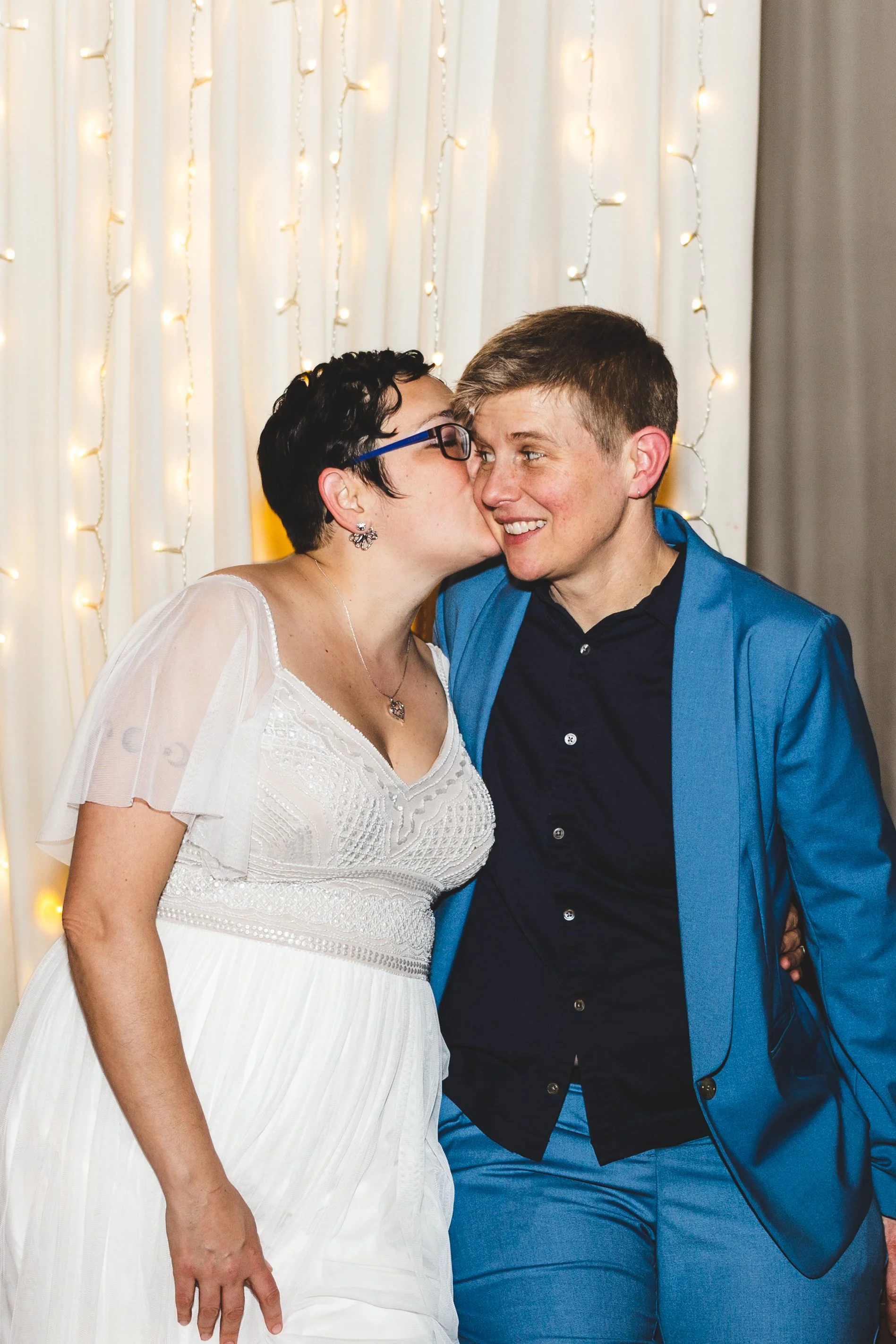 A woman in a white dress is giving a kiss on the cheek to a man in a blue suit, both smiling, in front of a backdrop of string lights and white curtains.