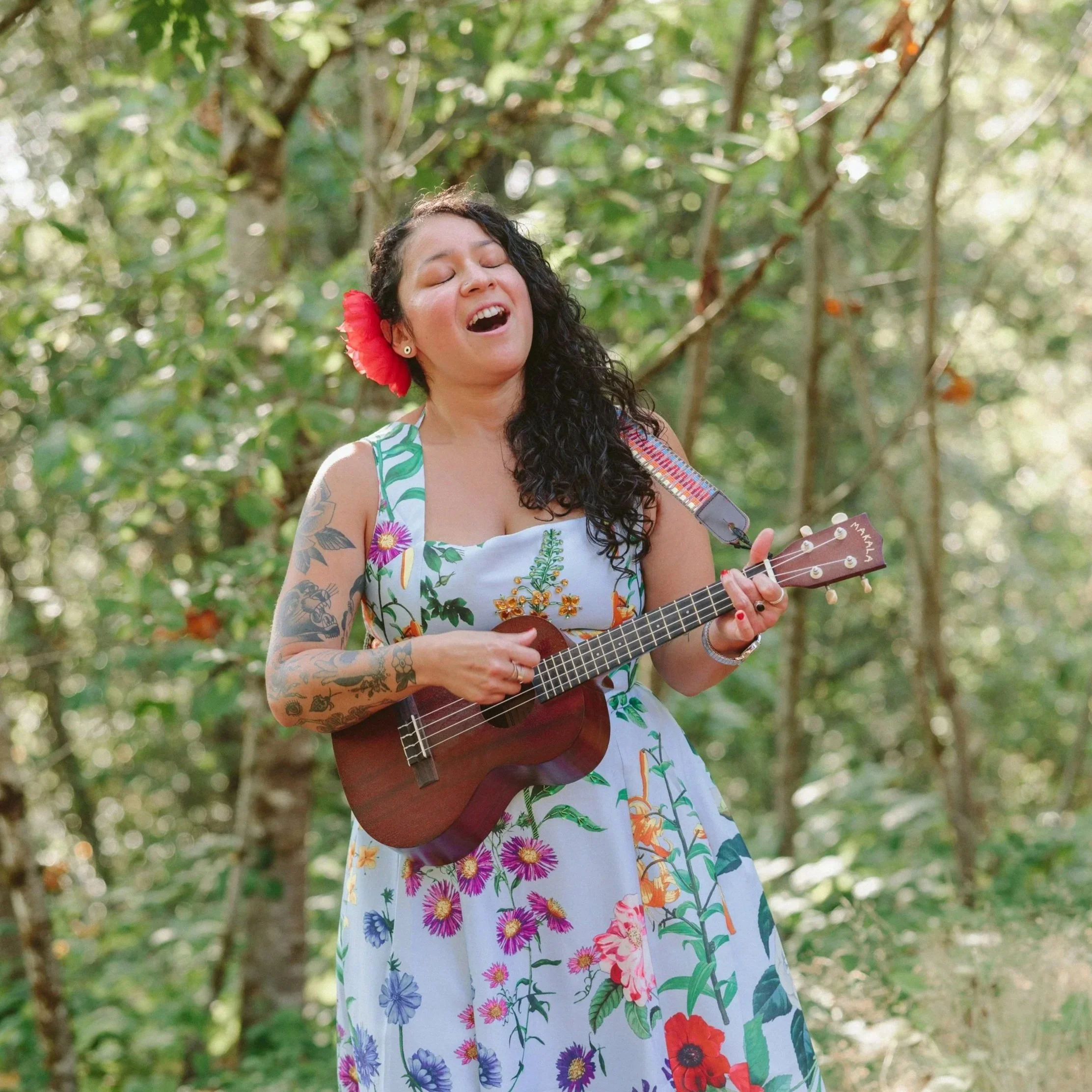 A woman with curly dark hair, wearing a white dress with colorful floral patterns, singing or playing a ukulele outdoors in a green, leafy setting.