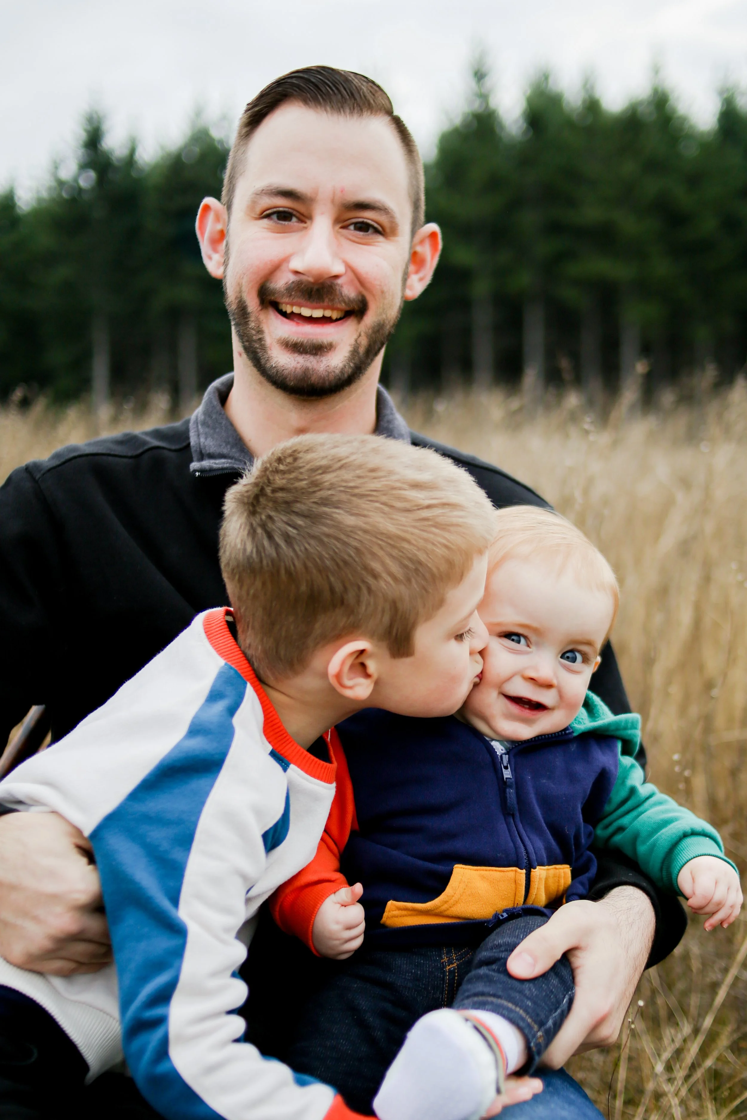 A smiling man holding two young children outdoors in a field with trees in the background.