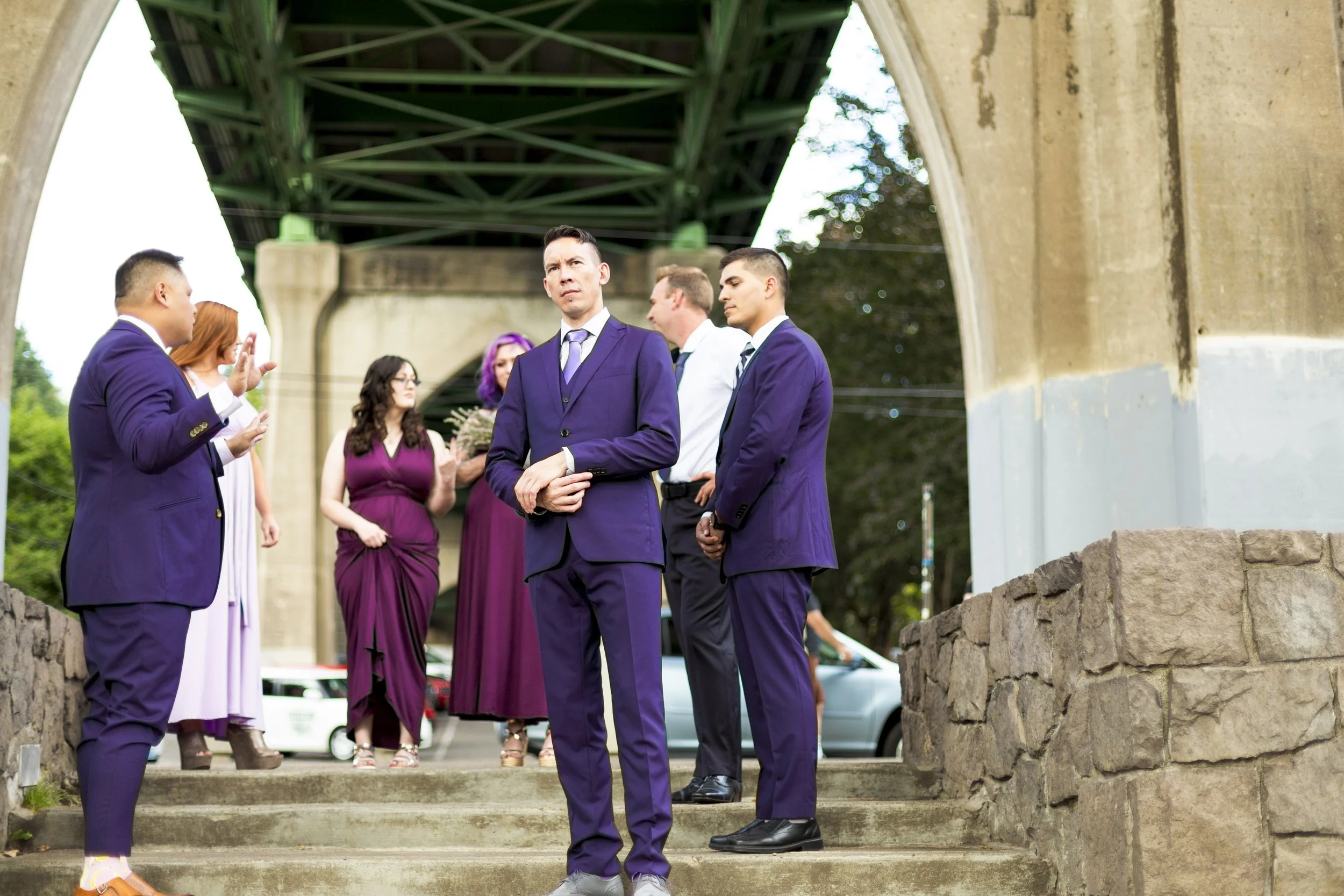 Group of people in formal attire standing on steps under a bridge, engaged in conversation.