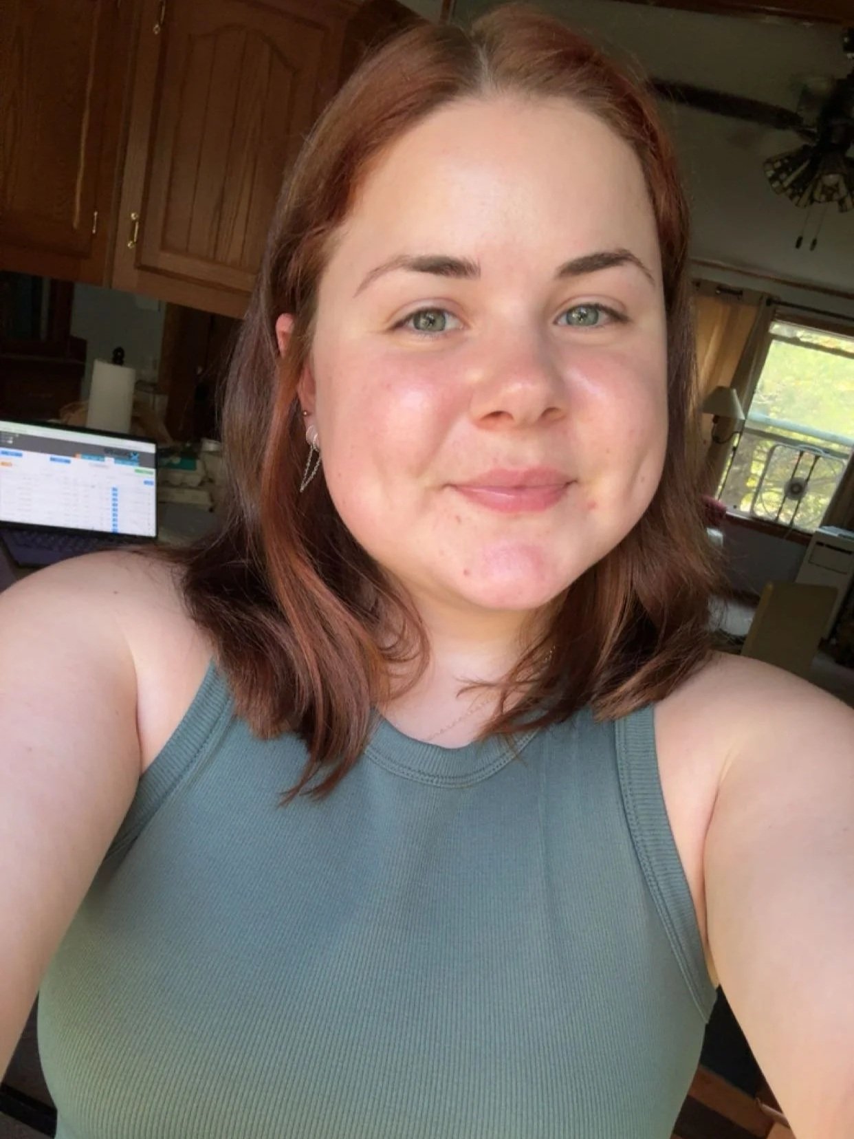 A woman with shoulder-length reddish-brown hair taking a selfie in a room with wooden cabinets and a ceiling fan. She's wearing a sleeveless gray top. In the background, a computer screen is visible with a spreadsheet open, and a window with curtains lets in natural light.