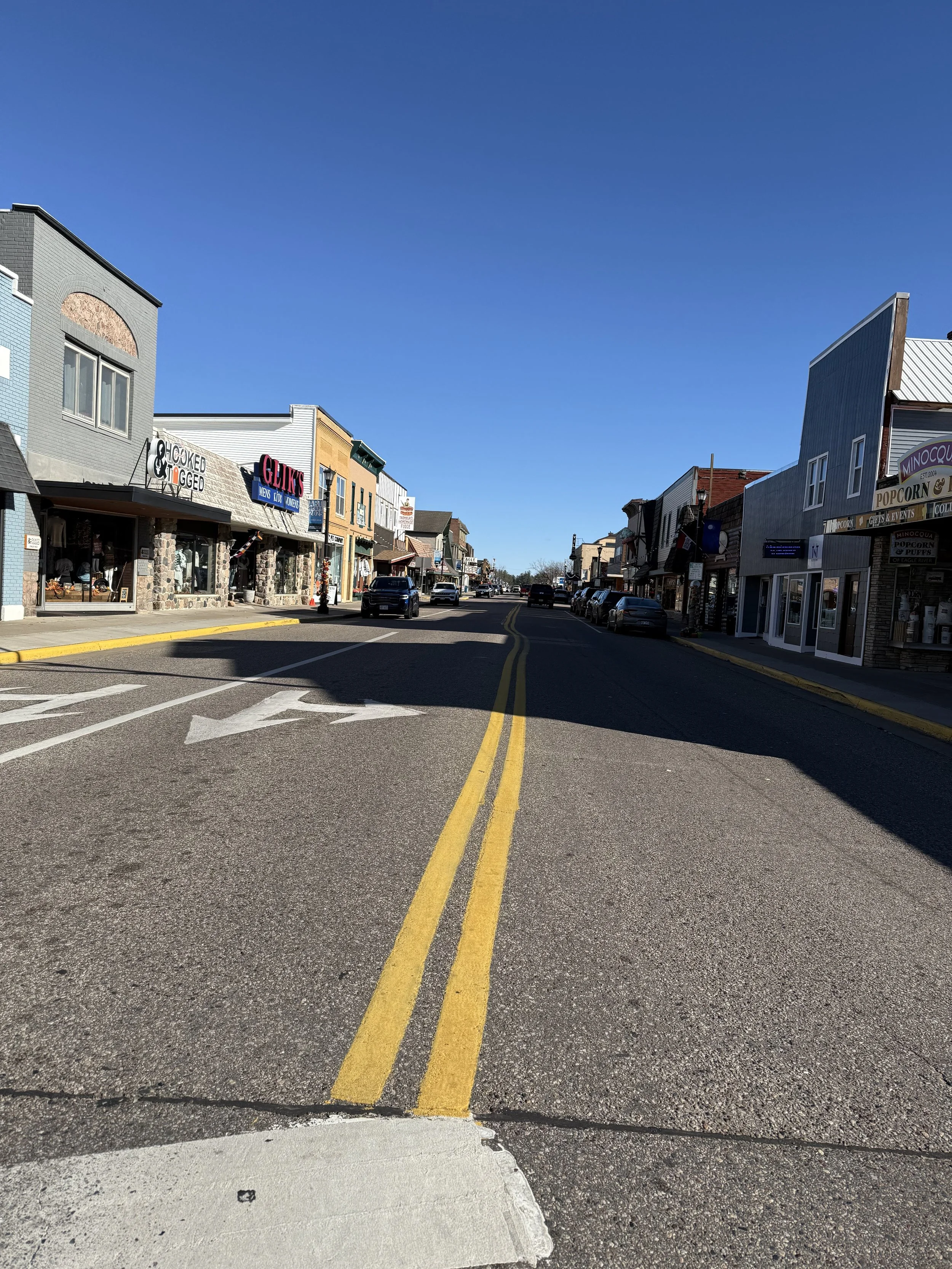 A small town street with storefronts on both sides, parked cars, and a clear blue sky.