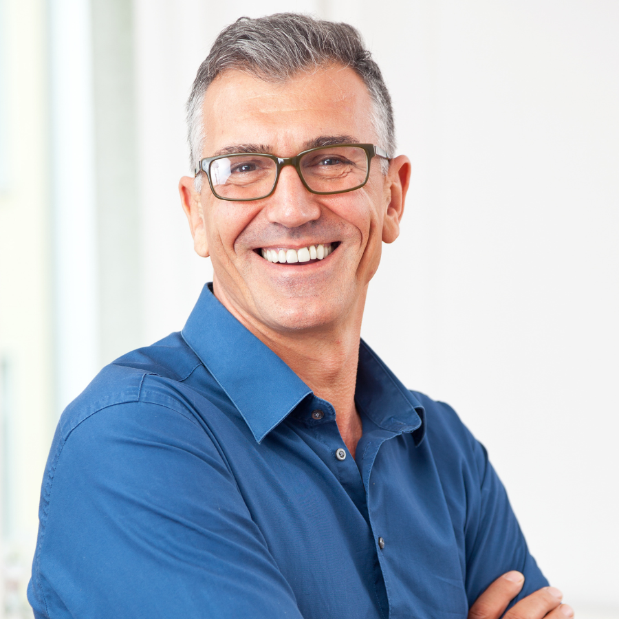 A smiling middle-aged man with gray hair, glasses, and a blue collared shirt standing with arms crossed in a well-lit room.