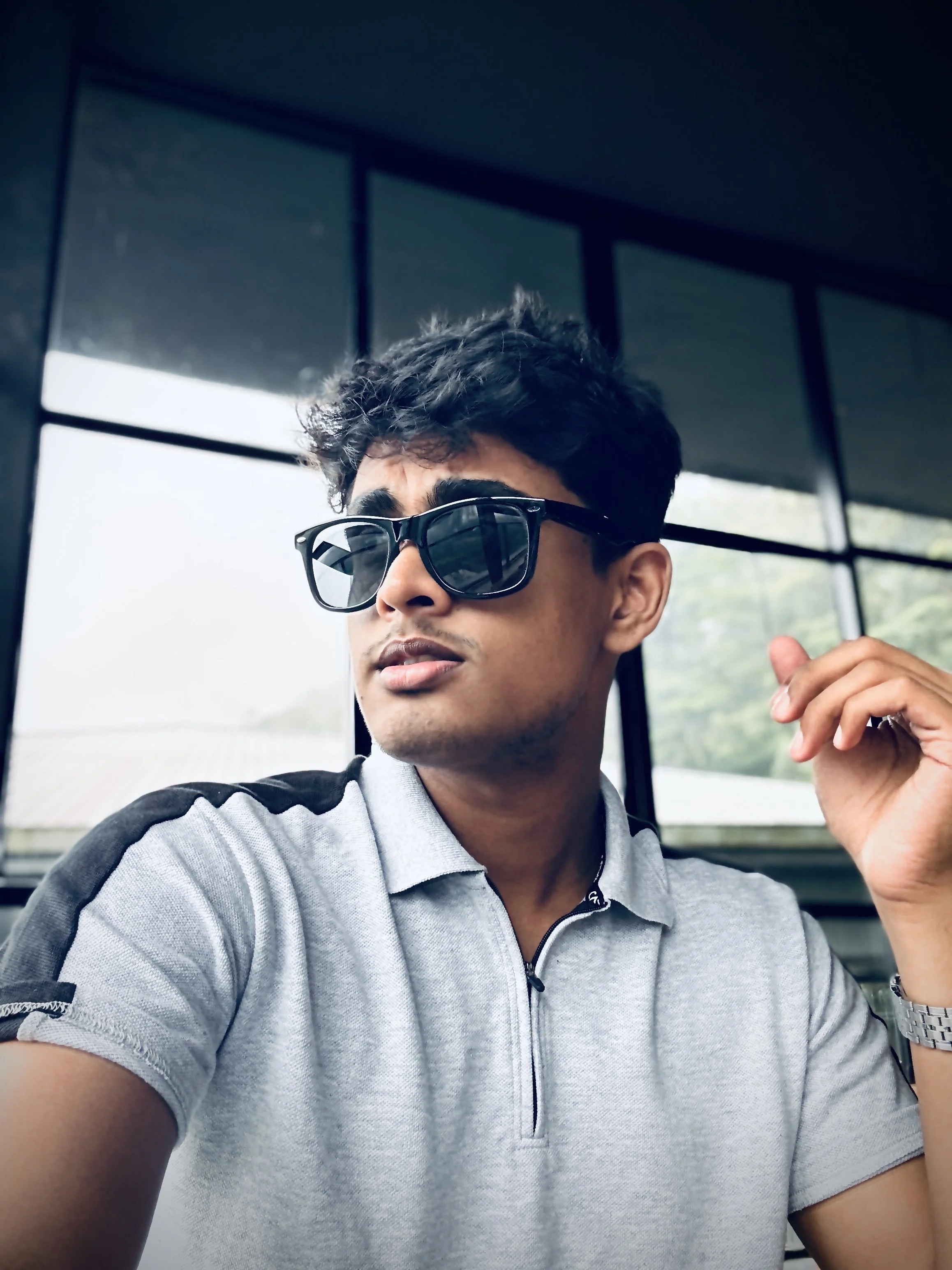 A young man wearing sunglasses and a light gray polo shirt, sitting indoors near large windows with a green outdoor view.