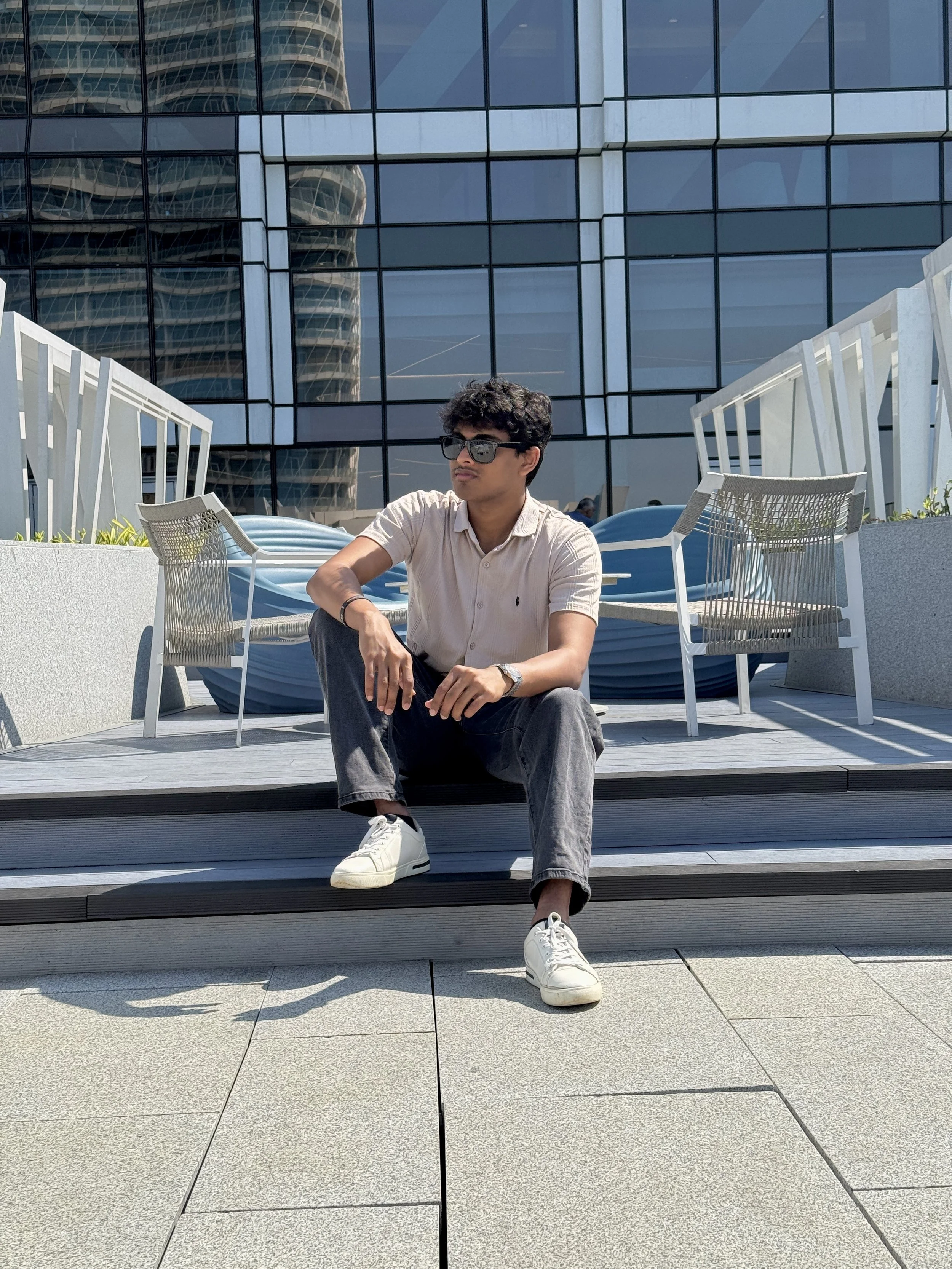 A young man with curly dark hair wearing sunglasses, a beige short-sleeve shirt, black pants, and white sneakers sitting on outdoor steps in front of a modern glass building with reflections of another high-rise.