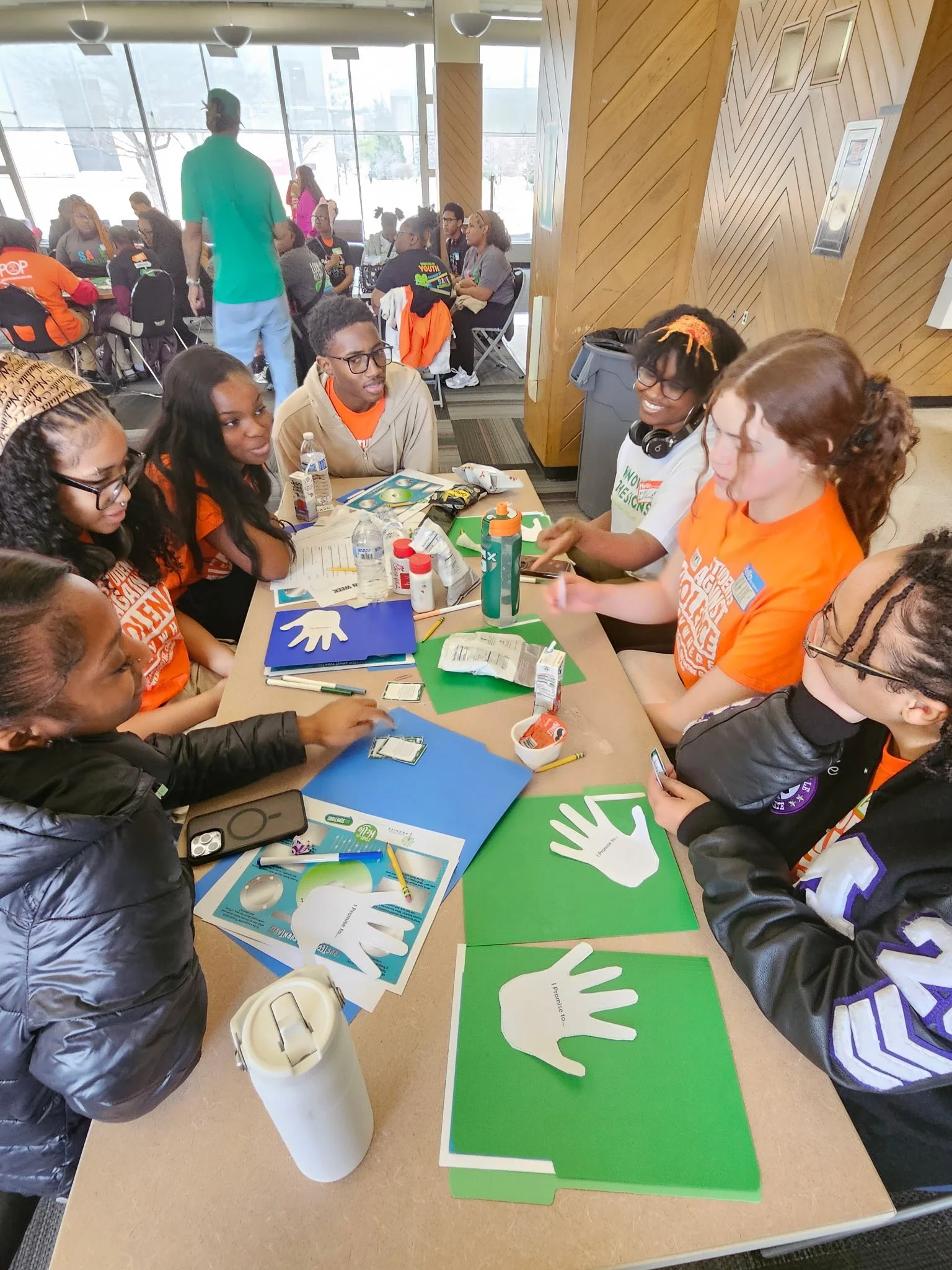 A group of people sitting around a table participating in a workshop or event, with various supplies such as paper cutouts shaped like hands, markers, and bottles on the table, and a large window in the background.
