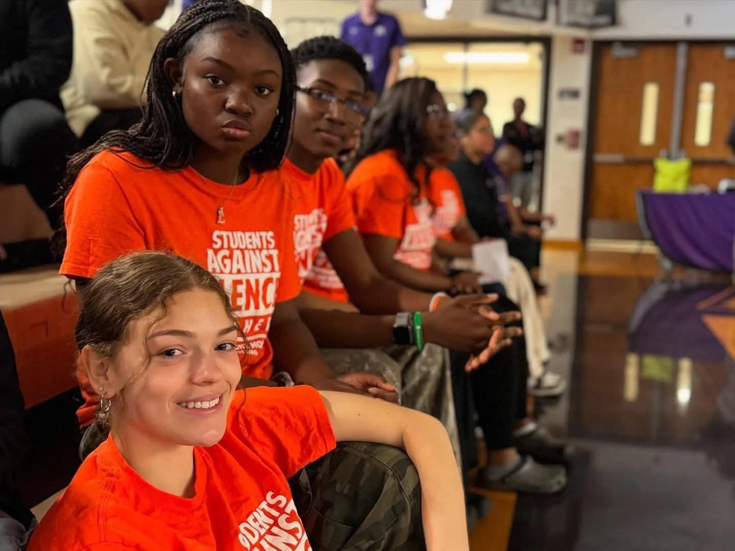 Group of young people sitting in a row, wearing orange t-shirts with text supporting a cause, in an indoor setting with wooden doors and walls.