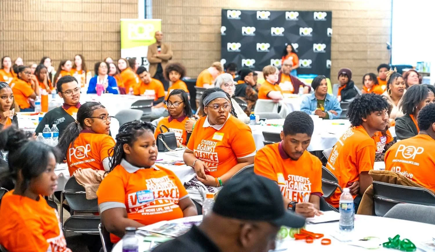 A large group of young people attending a conference, many wearing orange T-shirts with the phrase "Students Against Violence" printed on them. They are seated at round tables in a spacious room, listening attentively. Some have water bottles and papers on the tables. In the background, two people are standing near a banner and a screen.