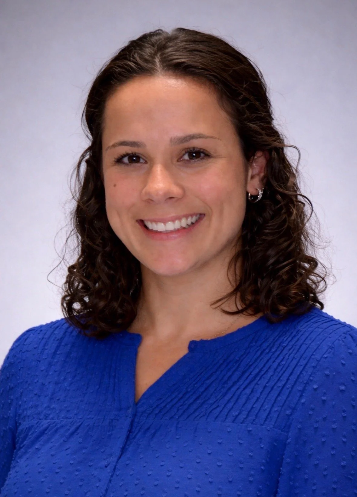 A woman with shoulder-length curly brown hair, wearing a textured blue top, smiling in front of a plain, light-colored background.