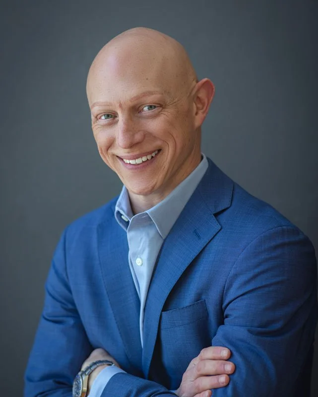 A smiling man with a bald head wearing a blue suit and light blue dress shirt, standing against a gray background.