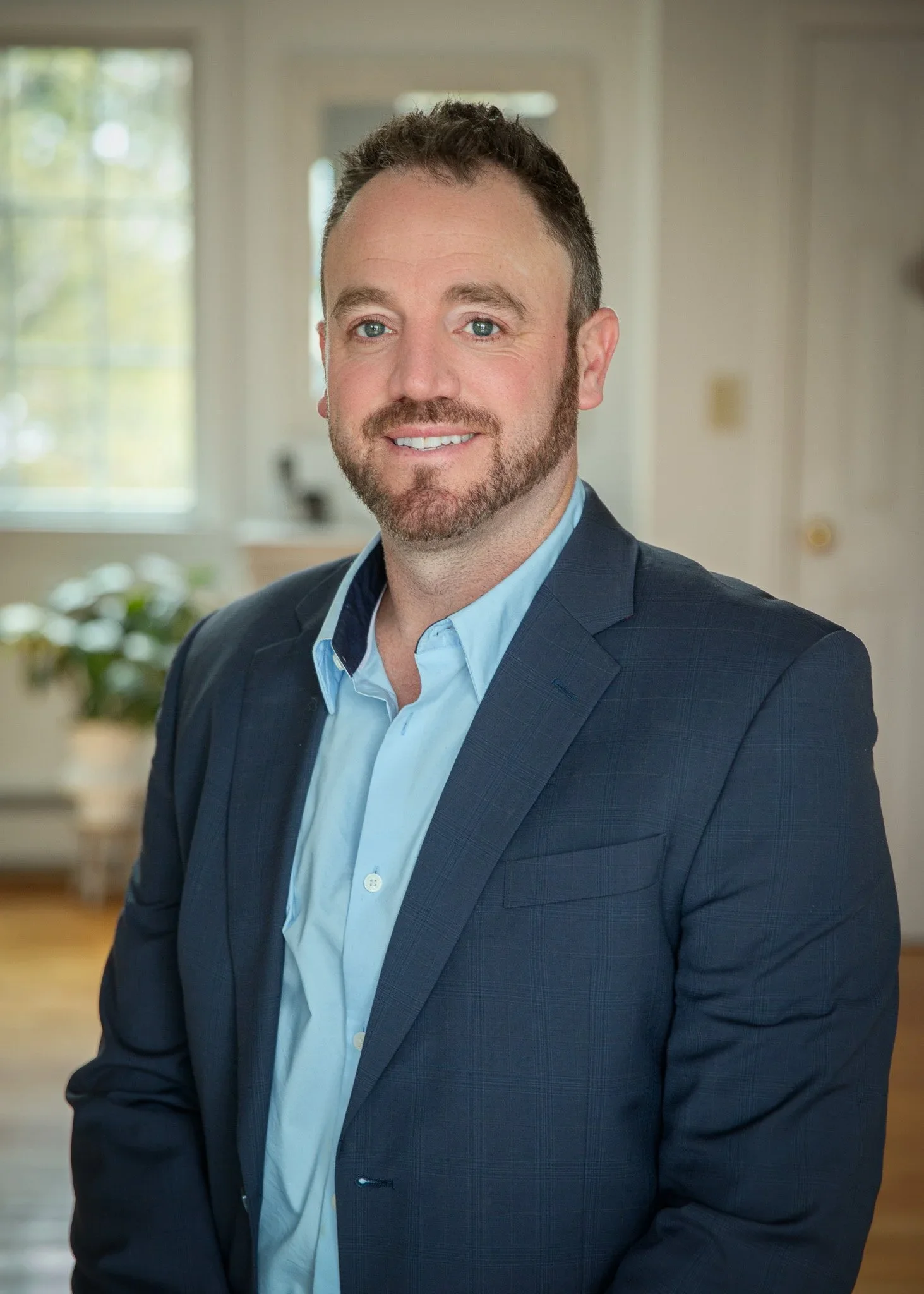 A man with short brown hair and a beard, wearing a dark suit jacket and a light blue button-up shirt, smiling in an indoor setting with blurred windows and greenery in the background.