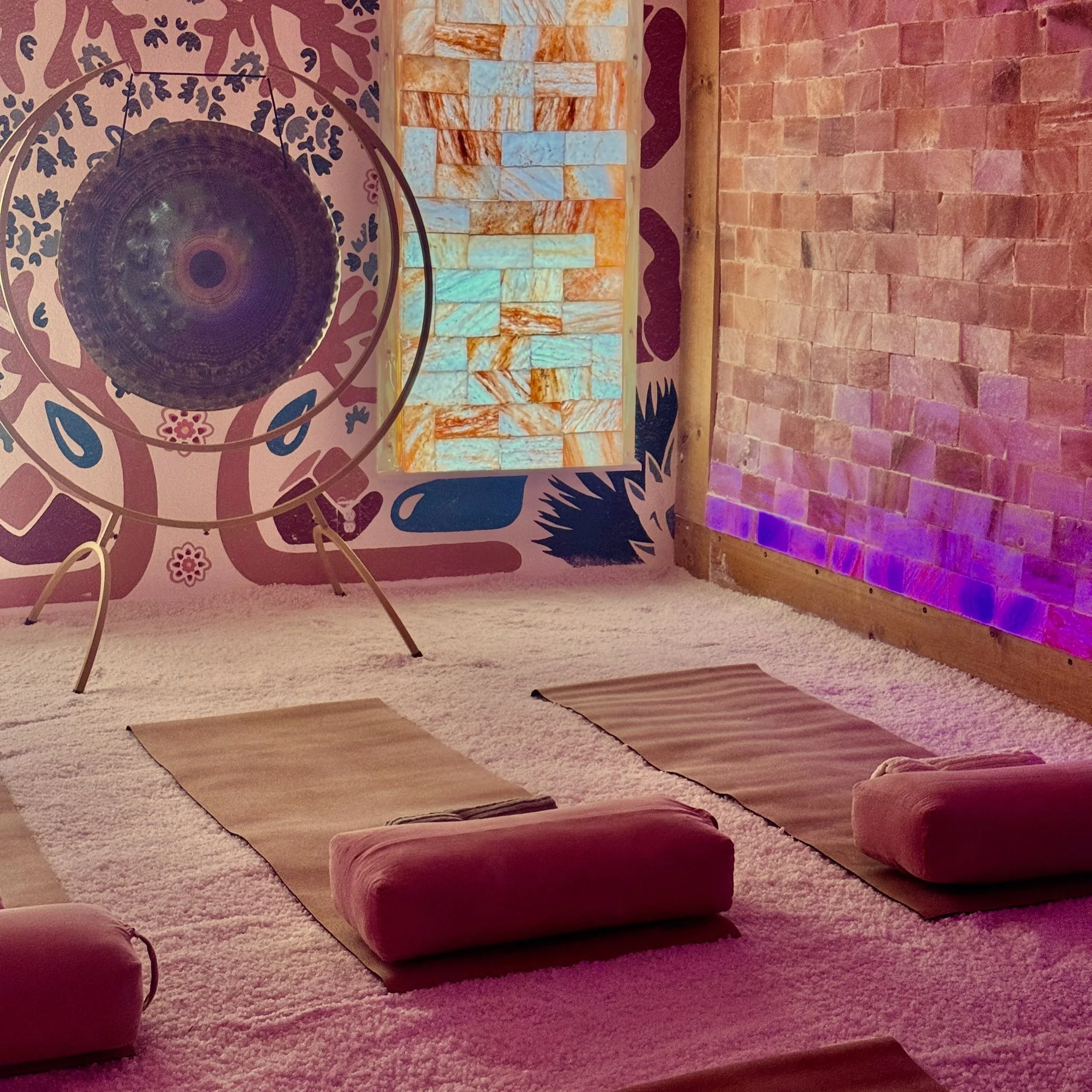 A room with a brick wall, purple colored wallpaper with leaf and flower patterns, a spherical metallic hanging light fixture, a row of colorful stone blocks on a wooden shelf, several yoga mats and cushions on a beige carpet, and some blue LED strip lights along the bottom of the brick wall.