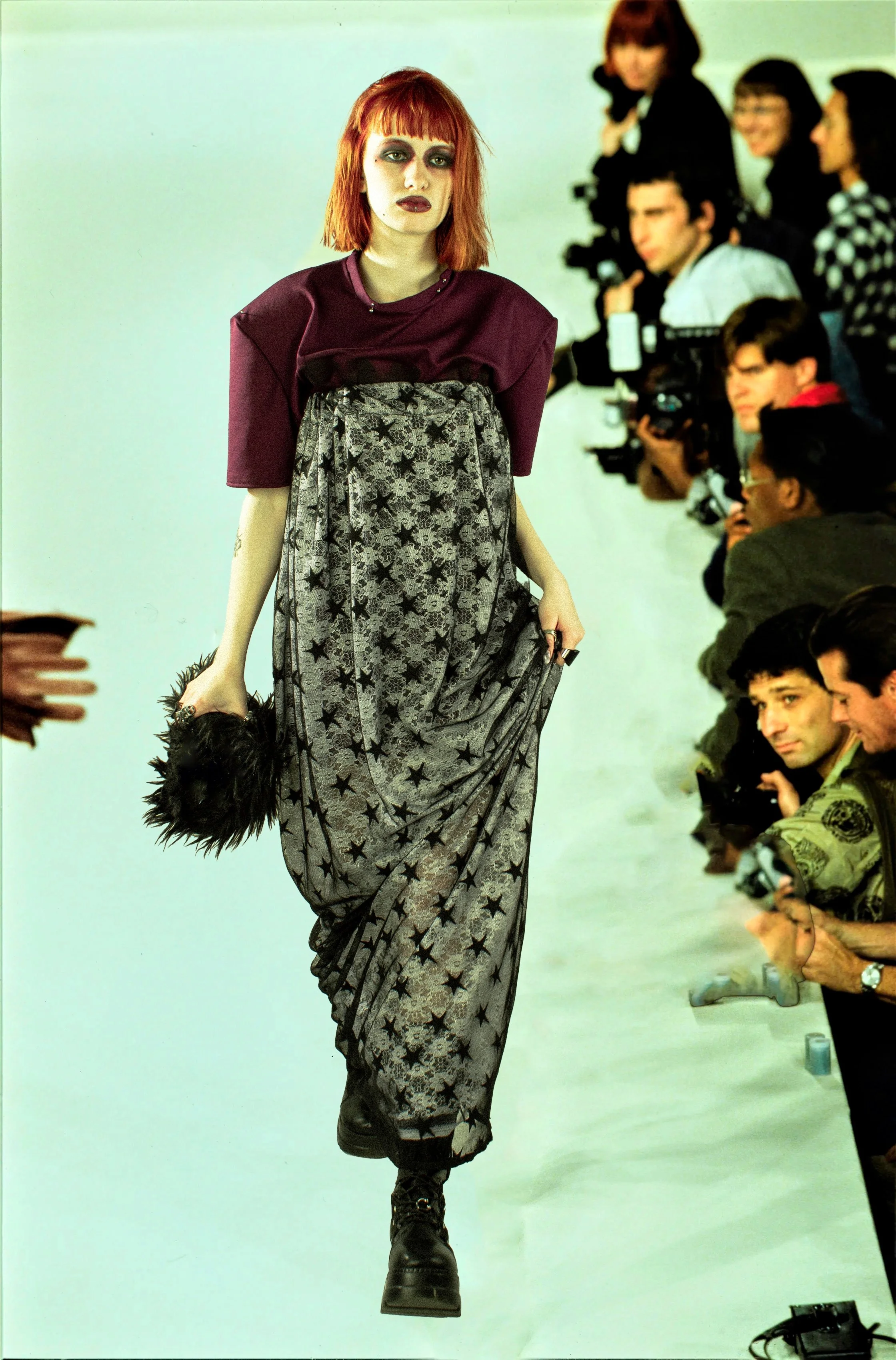 Modelo caminando en una pasarela con espectadores tomando fotos, lleva un vestido largo con estampado de estrellas y botas negras, cabello rojo y maquillaje oscuro.