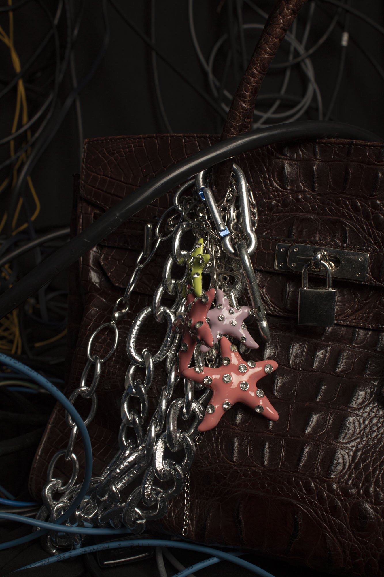 Un bolso de color marrón oscuro con textura de cuero de cocodrilo y candado, accesorios de llaves y collares colgados de la manija, rodeado de cables y cables eléctricos en un entorno oscuro.