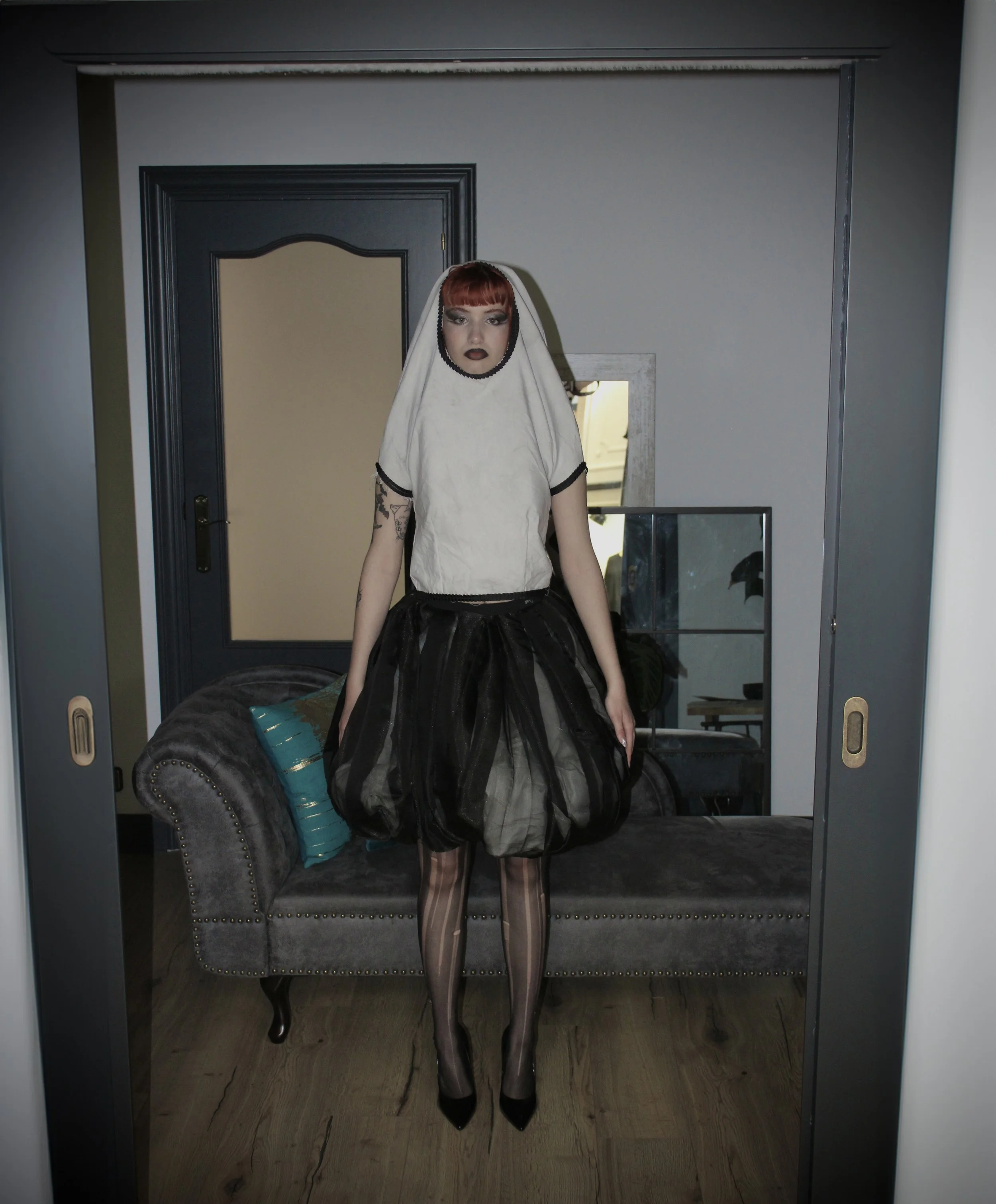 Persona con cabello rojo, maquillaje oscuro y vestido de malla y capa negra, de pie frente a un espejo en un entorno moderno y elegante.