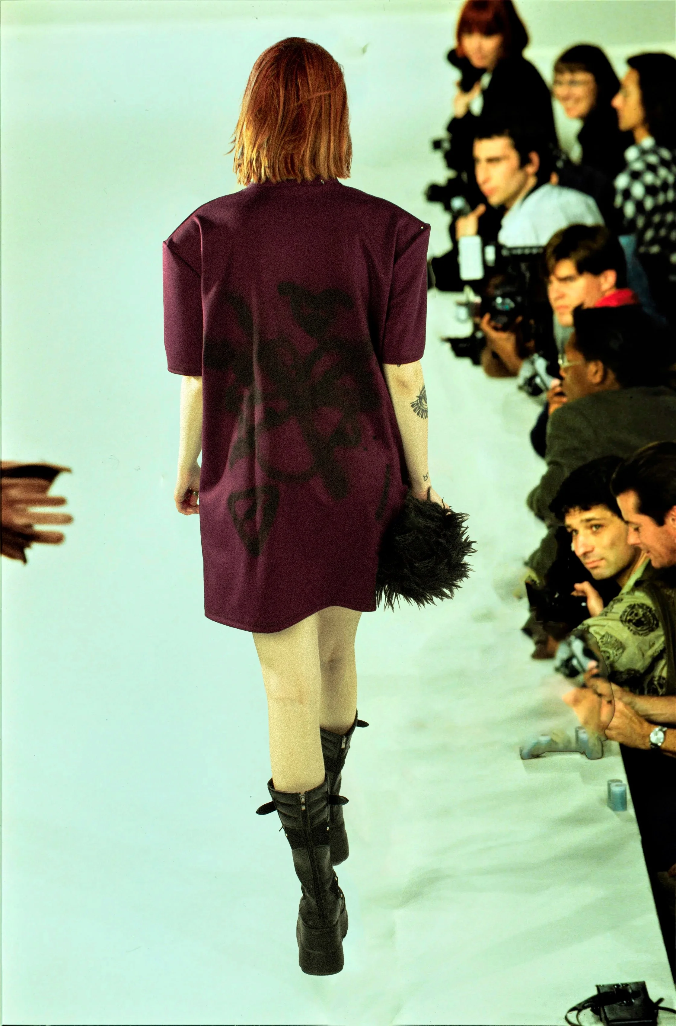 Mujer con cabello rubio y ropa morada, vista de espaldas, caminando en una pasarela durante un evento de moda, rodeada de fotógrafos y espectadores.