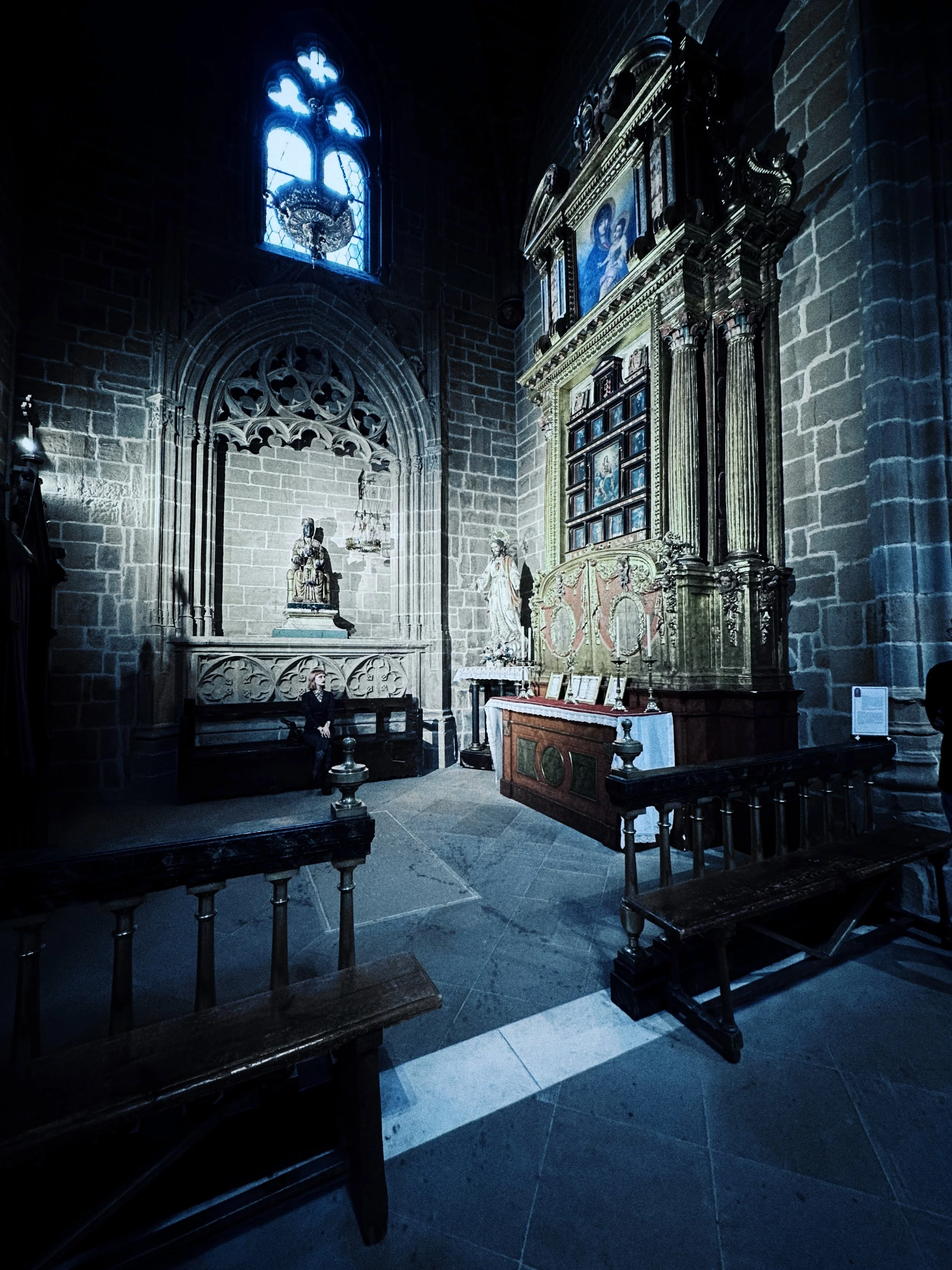 Interior de una iglesia con vitrales azules y altares, bancos de madera y figuras religiosas. La luz de los vitrales ilumina el espacio en tonos azules y oscuros.