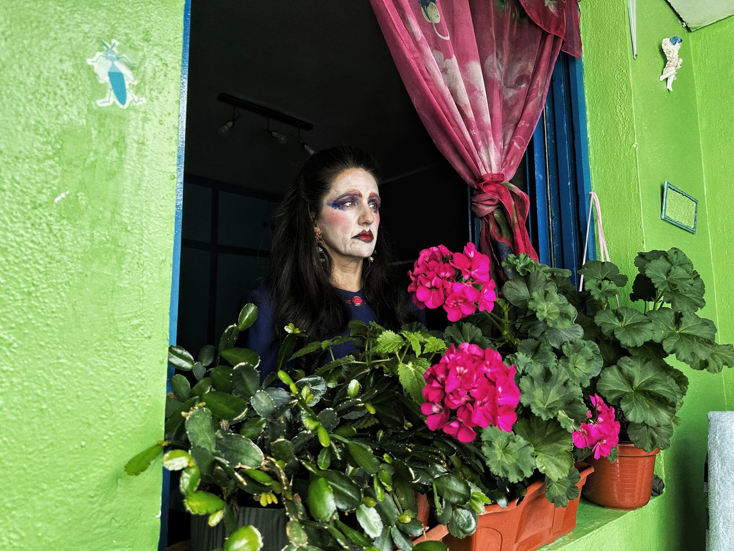 Mujer con maquillaje dramático mirando desde un balcón decorado con plantas y flores en macetas, con paredes verdes y cortinas rosas.