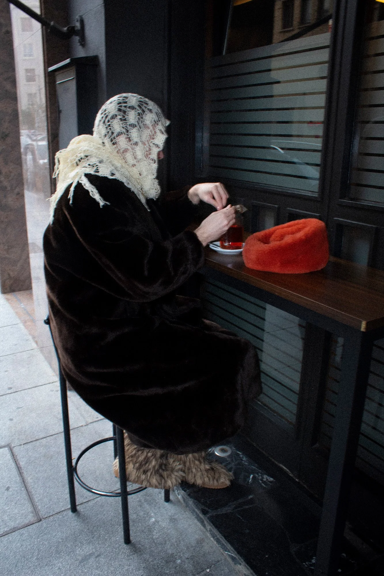 Una mujer con un pañuelo de encaje en la cabeza, sentada en una silla en un espacio interior, sirviendo una bebida en un vaso en una bandeja, con un bolso de peluche naranja sobre la mesa.