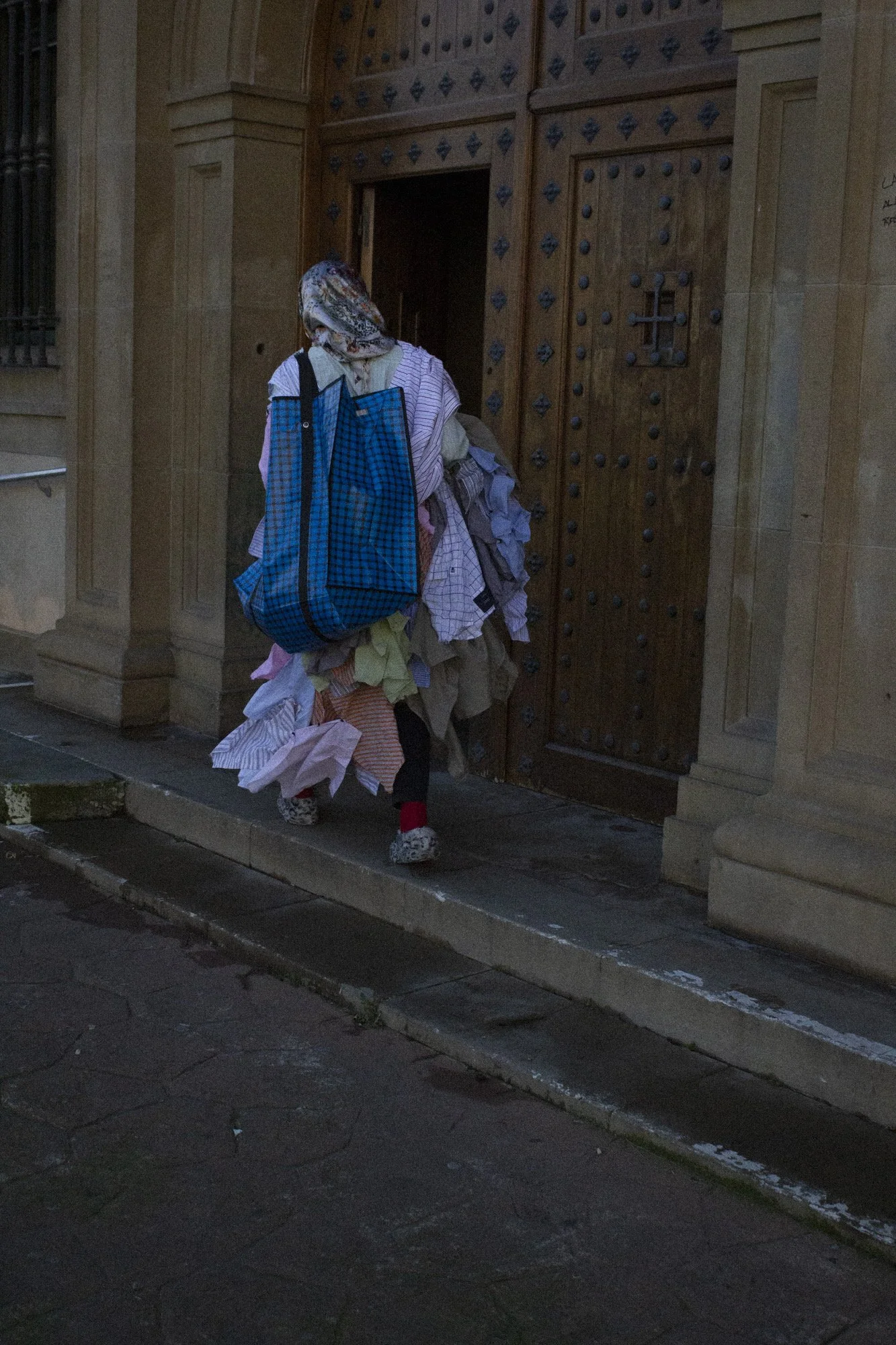 Persona con vestimenta colorida y saco de bolsas de tela, caminando por la acera frente a una puerta grande de madera con herrajes, en un entorno urbano por la noche.