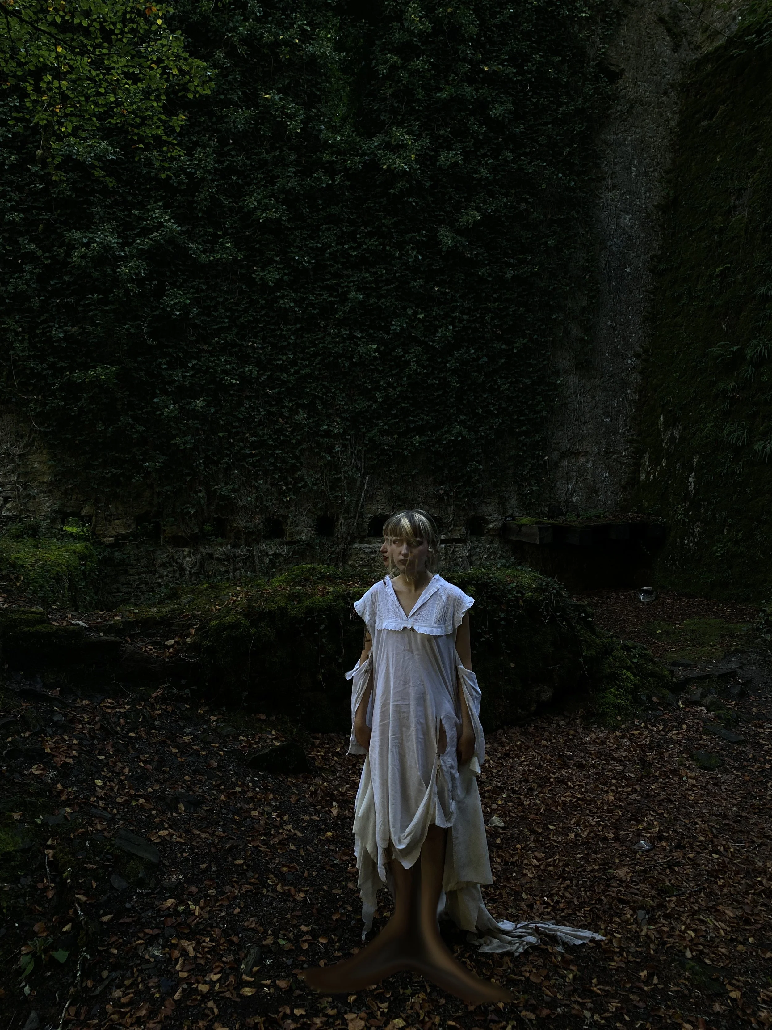Mujer con vestido largo de color blanco en un bosque oscuro con vegetación densa.