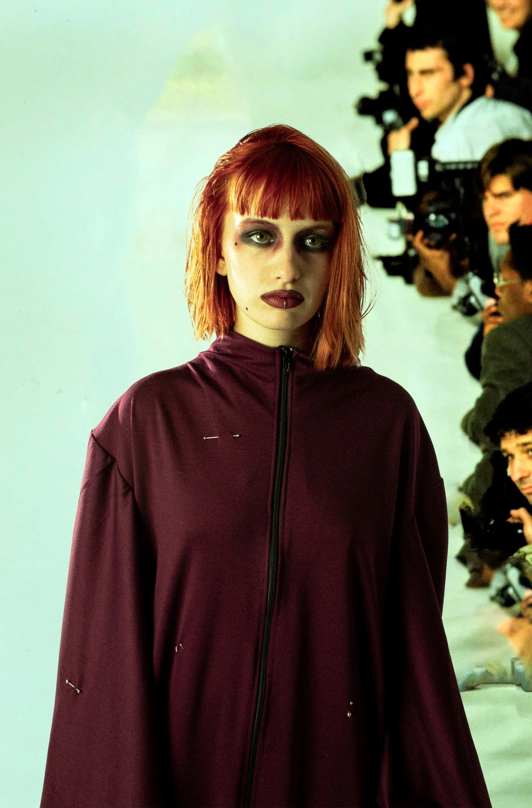 Mujer con cabello rojo, maquillaje oscuro y ropa de color burdeos en una pasarela con fotógrafos en el fondo.