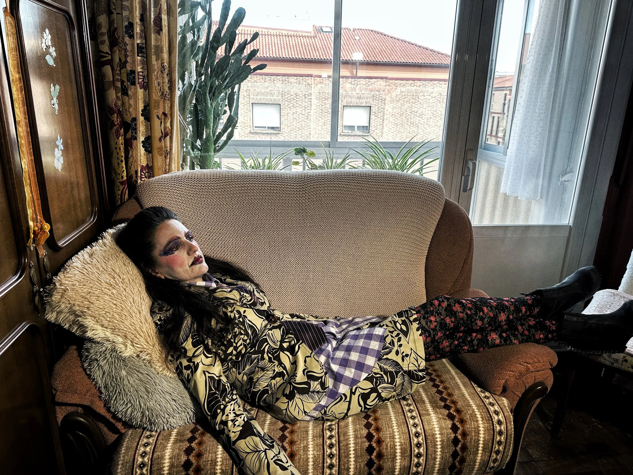 Una mujer con maquillaje extravagante y peinado oscuro recostada en un sofá en una habitación con ventanas grandes y cortinas, al fondo se ven edificios con techos rojos.